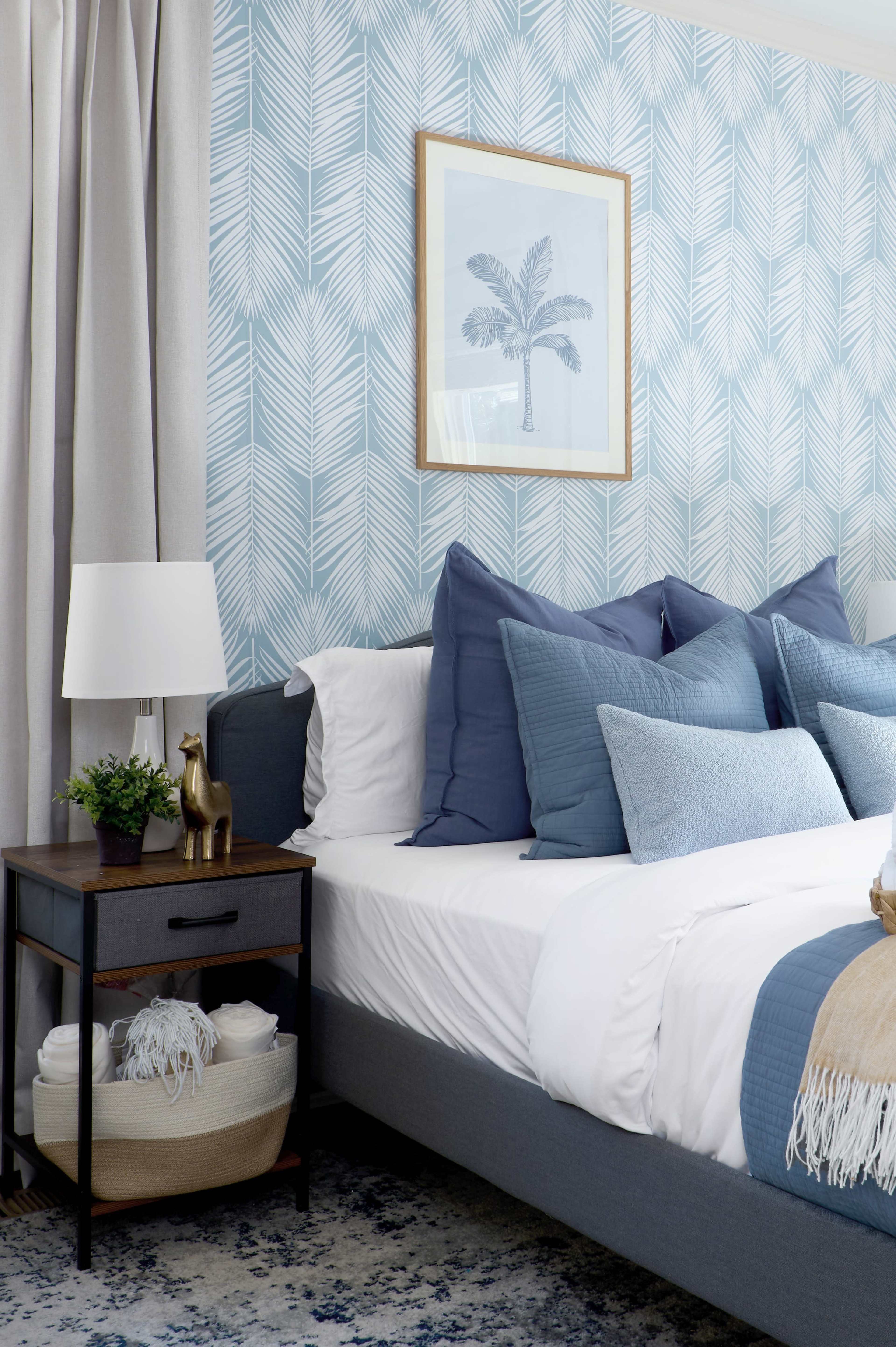 The image depicts a well-decorated bedroom featuring a bed with multiple blue pillows, a bedside table with a lamp, and a framed artwork of a palm tree against a patterned wallpaper.
