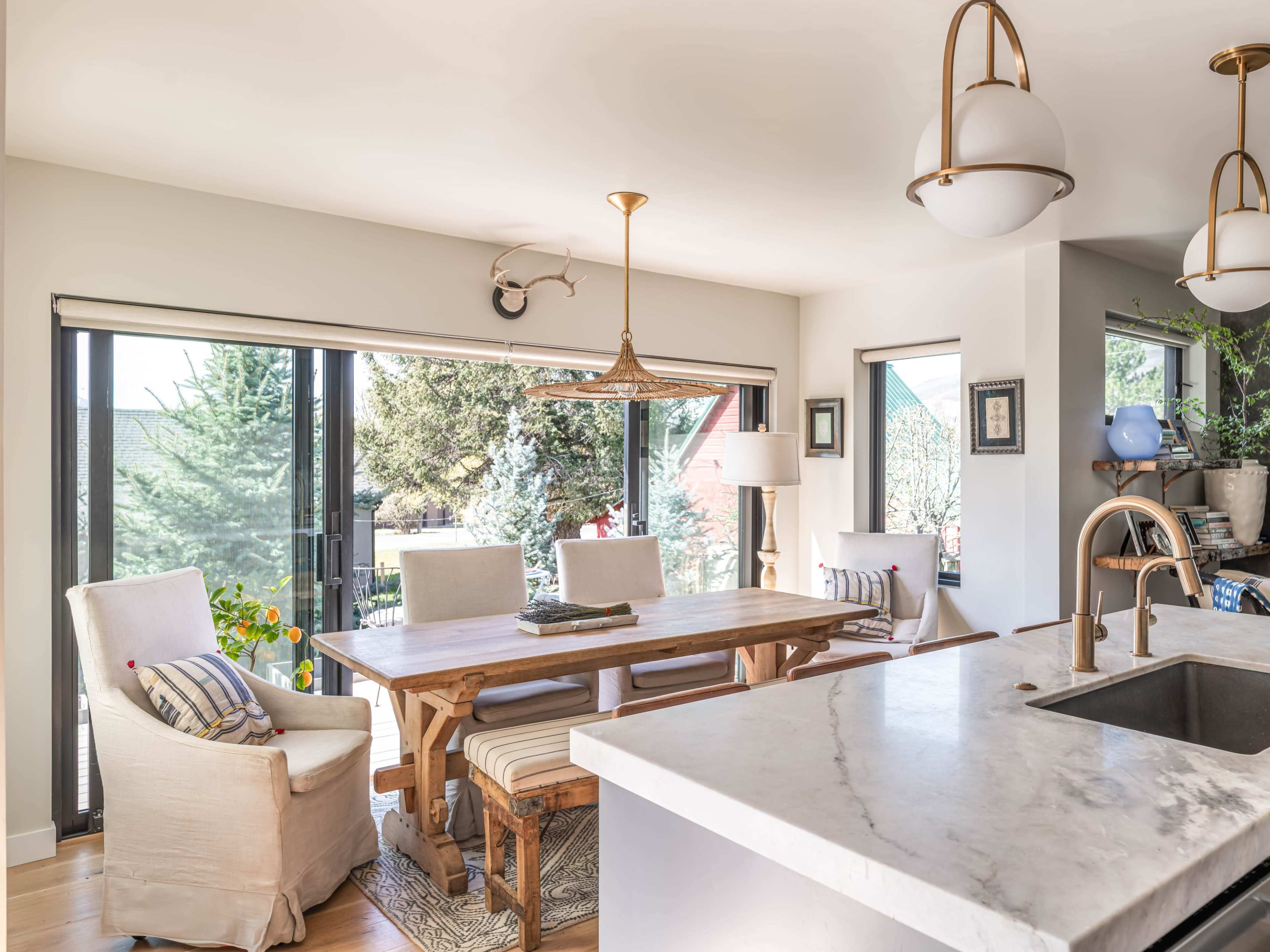 The image shows a modern dining area featuring a wooden table surrounded by upholstered chairs, large windows with outdoor views, and a kitchenette with a marble countertop.