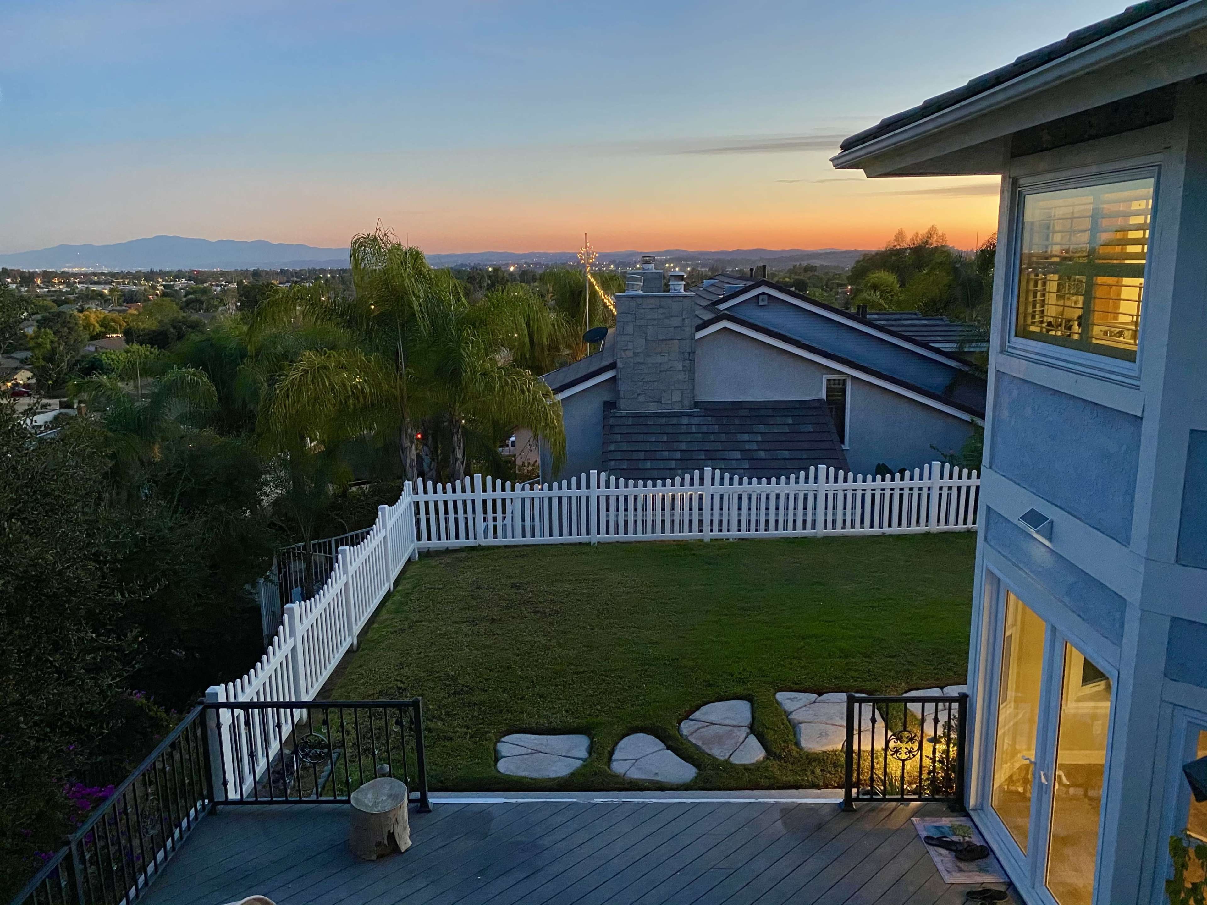 A hillside view from a house with a fenced backyard, overlooking a valley at sunset.