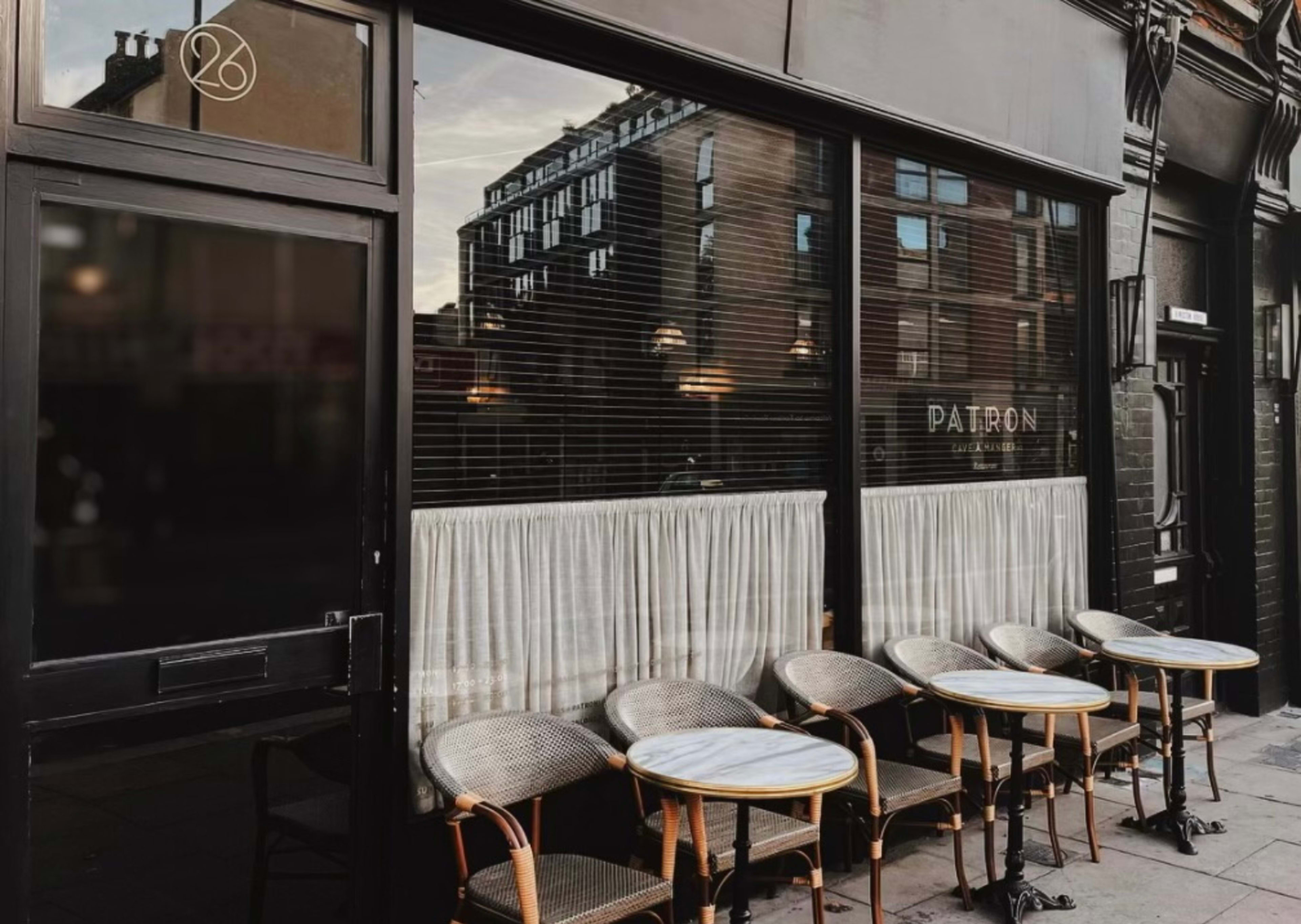 A café with several outdoor tables and chairs arranged in front of a large window displaying the word "PATRON" and white curtains.