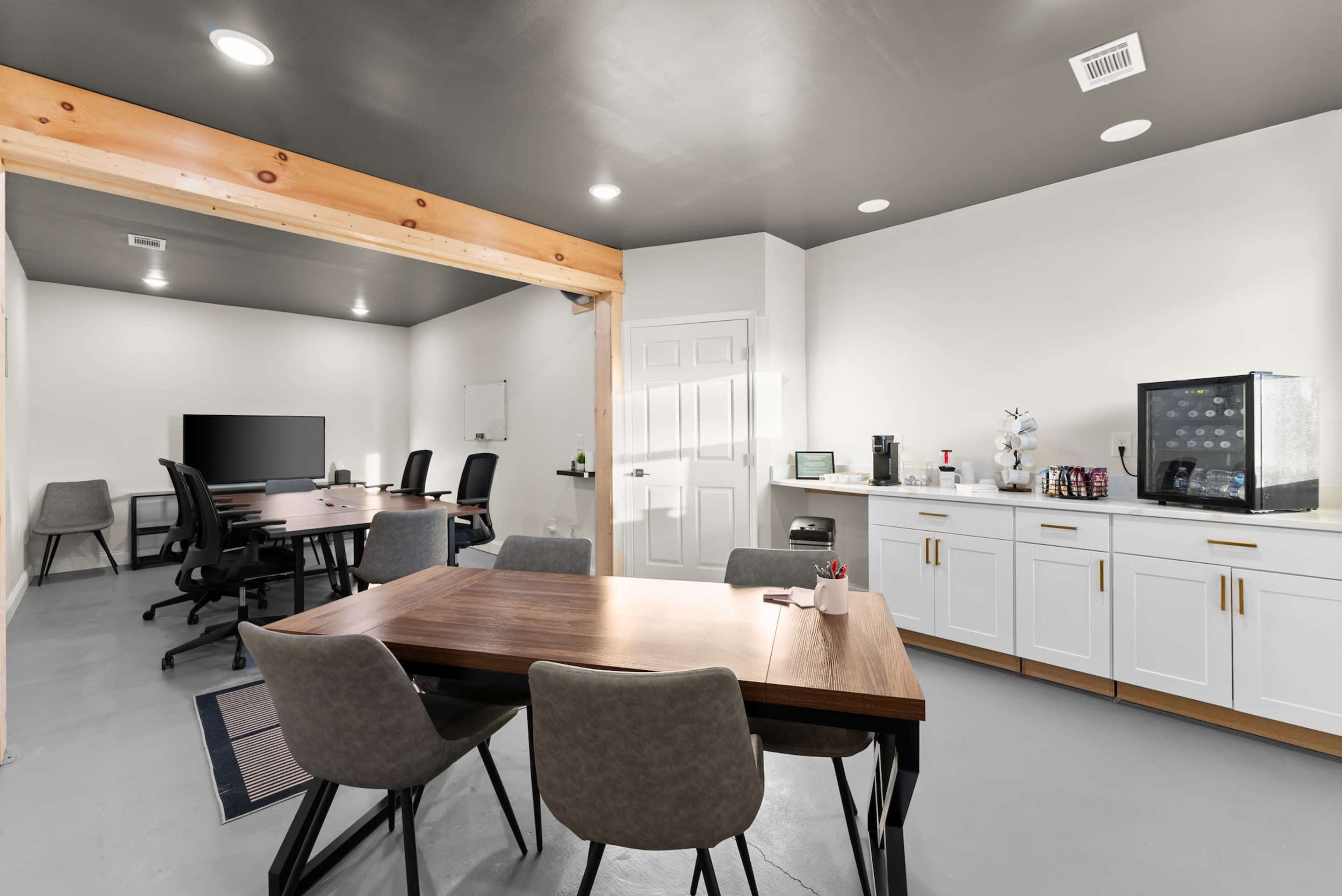 The image shows a modern office space featuring a meeting area with a large table and chairs, a separate workspace with a television, and a kitchenette equipped with appliances.