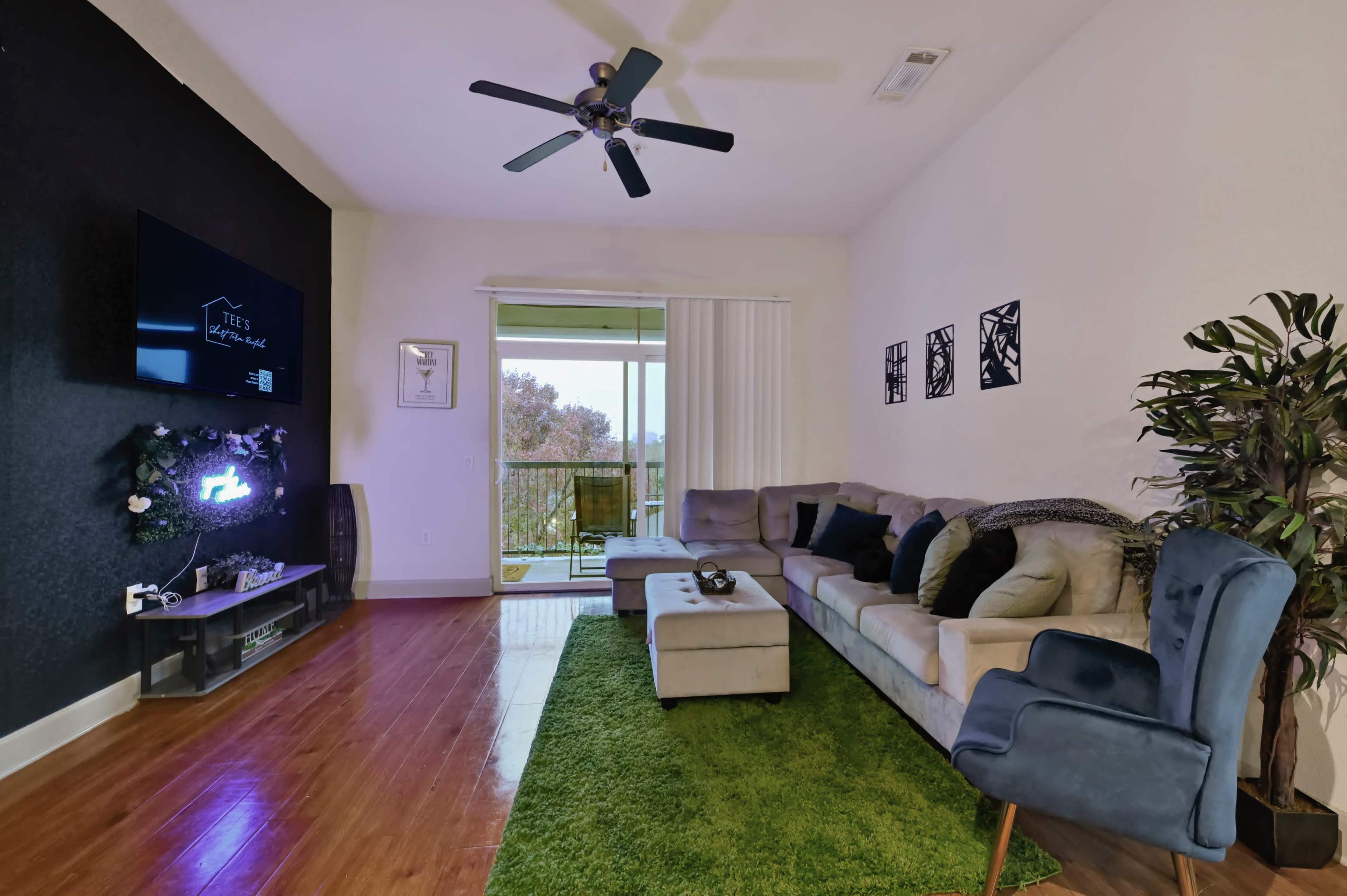 The living room features a sectional couch, a coffee table, a fan, and a large window that opens to a balcony with scenic views.
