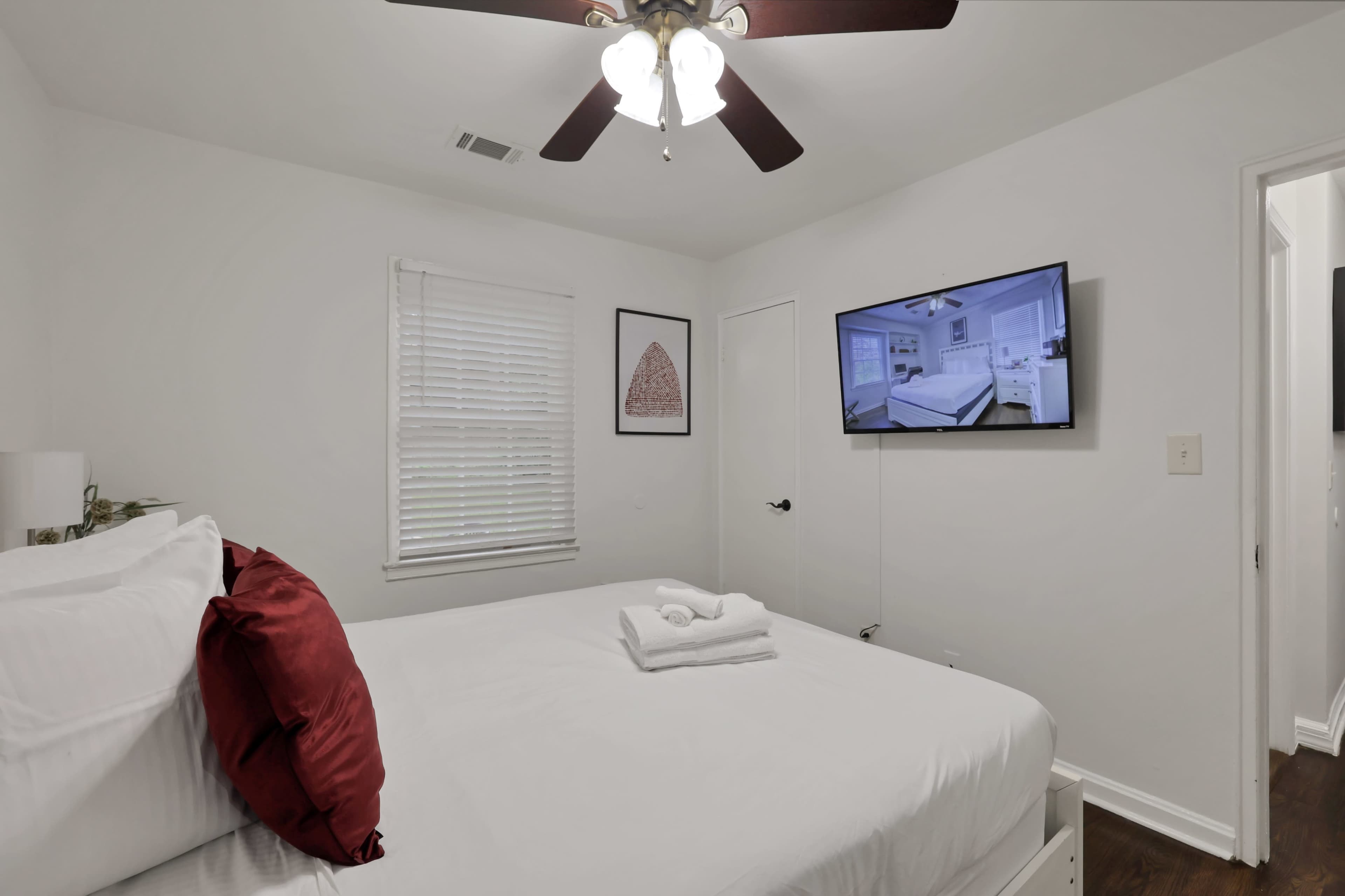 A small bedroom features a white bed with red pillows, a neatly arranged stack of towels, and a wall-mounted television.
