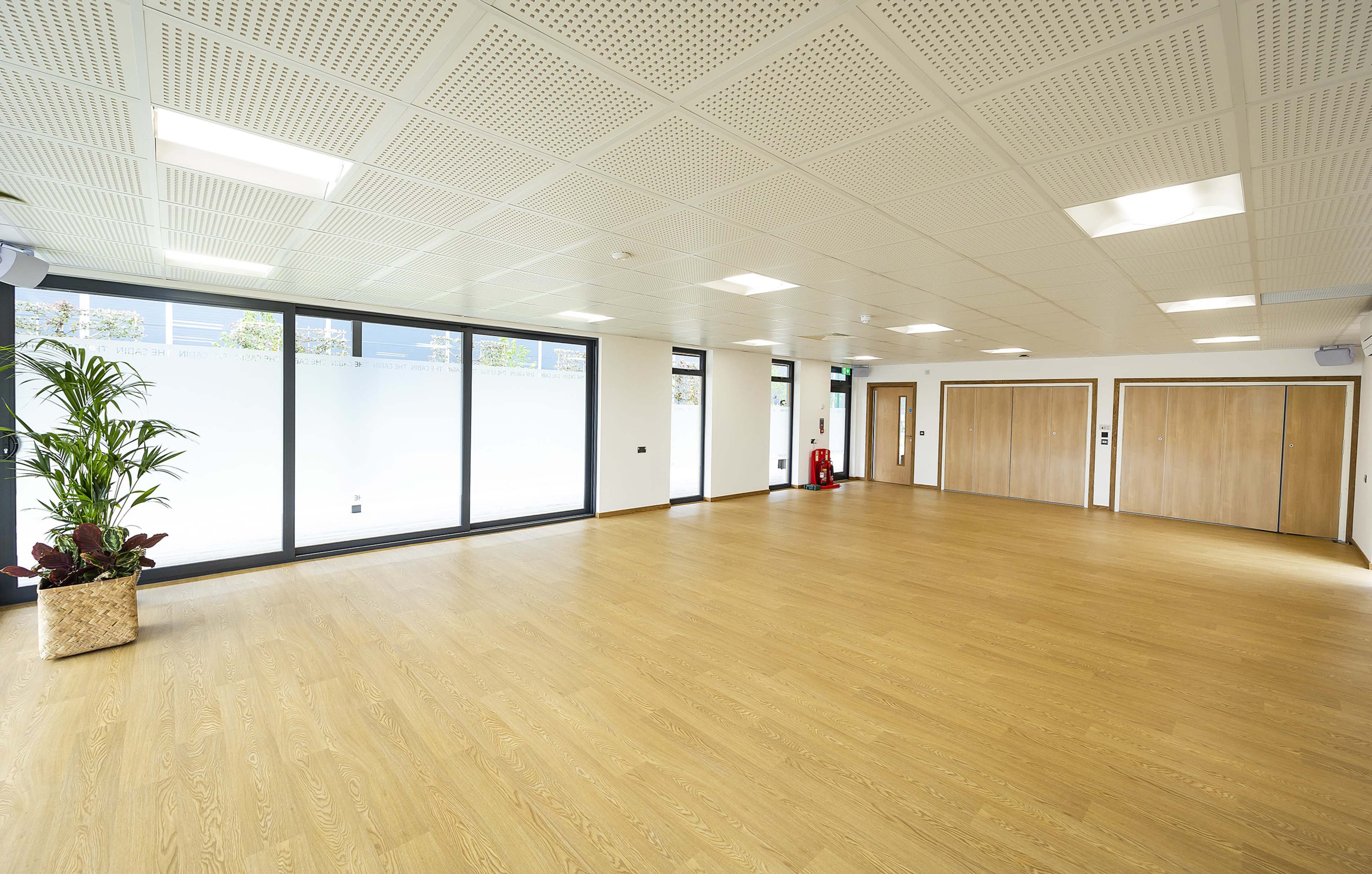 An empty indoor space with wooden flooring, large windows, and two closed doors on one side.