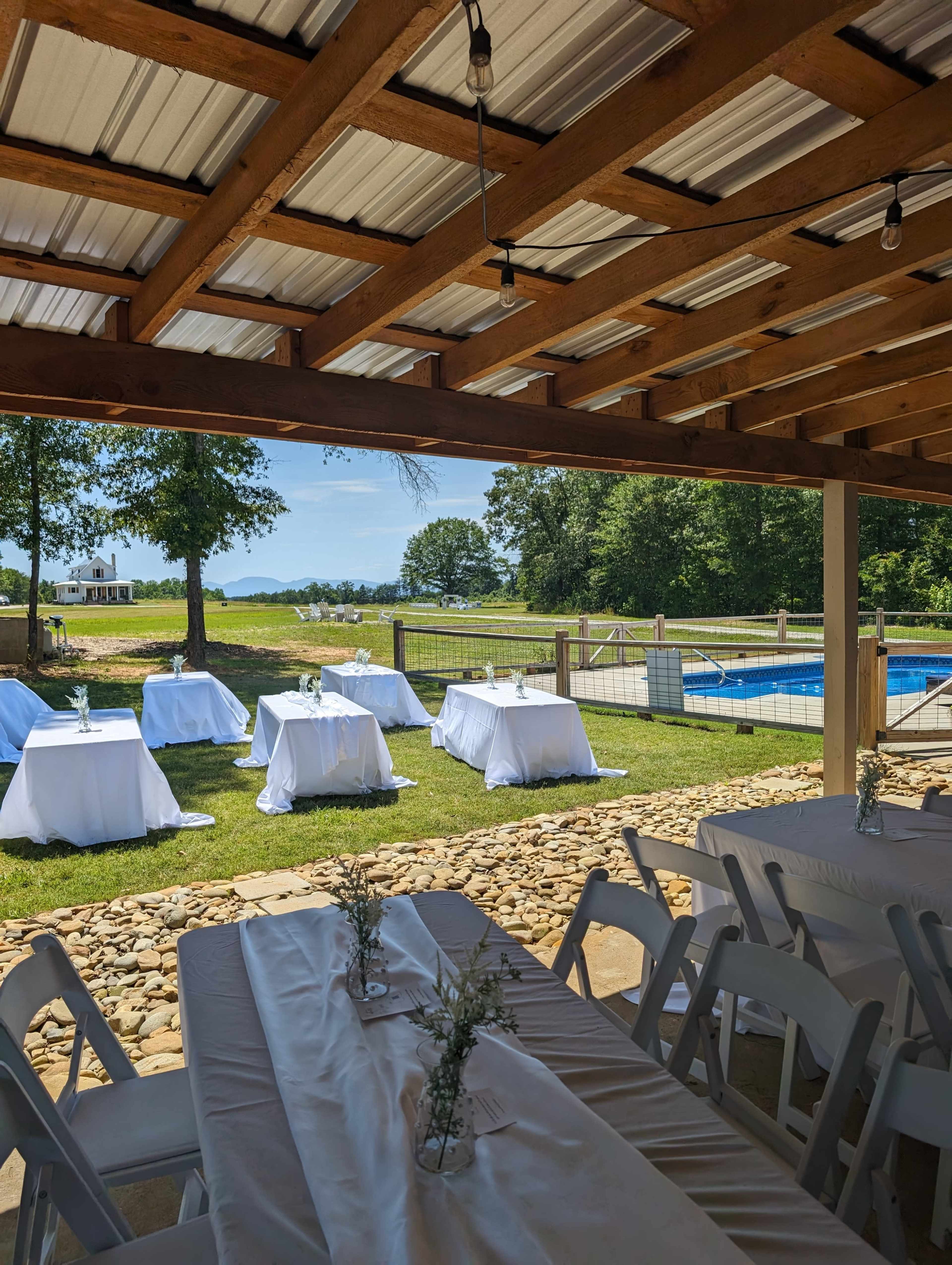 A spacious outdoor area with white-covered tables set up around a pool, surrounded by green grass and trees.