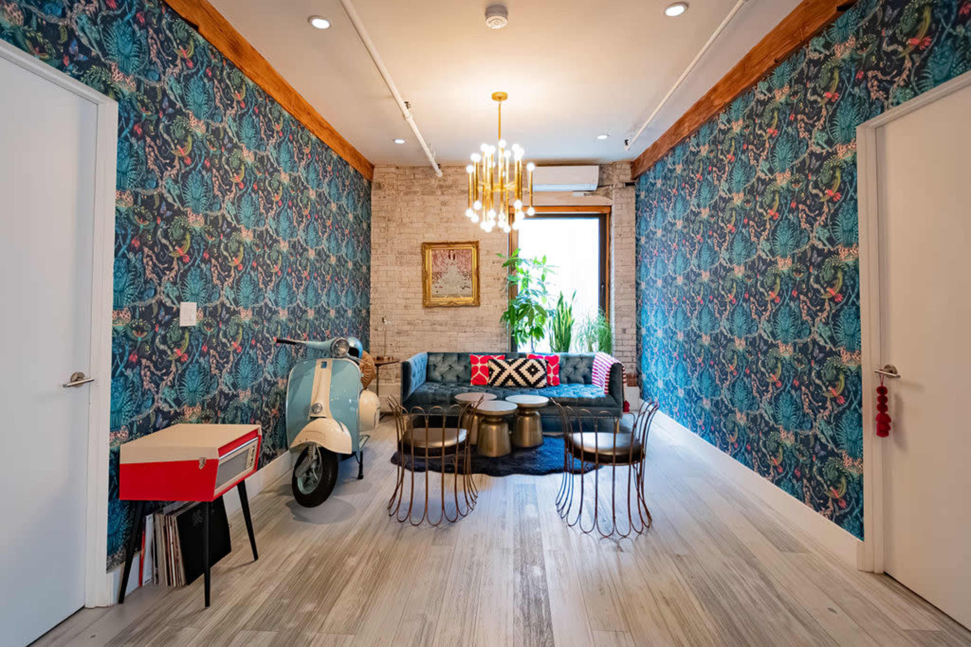 The image shows a modern living space with patterned wallpaper, a blue sofa, and a decorative plant, featuring a vintage scooter and unique metallic tables.