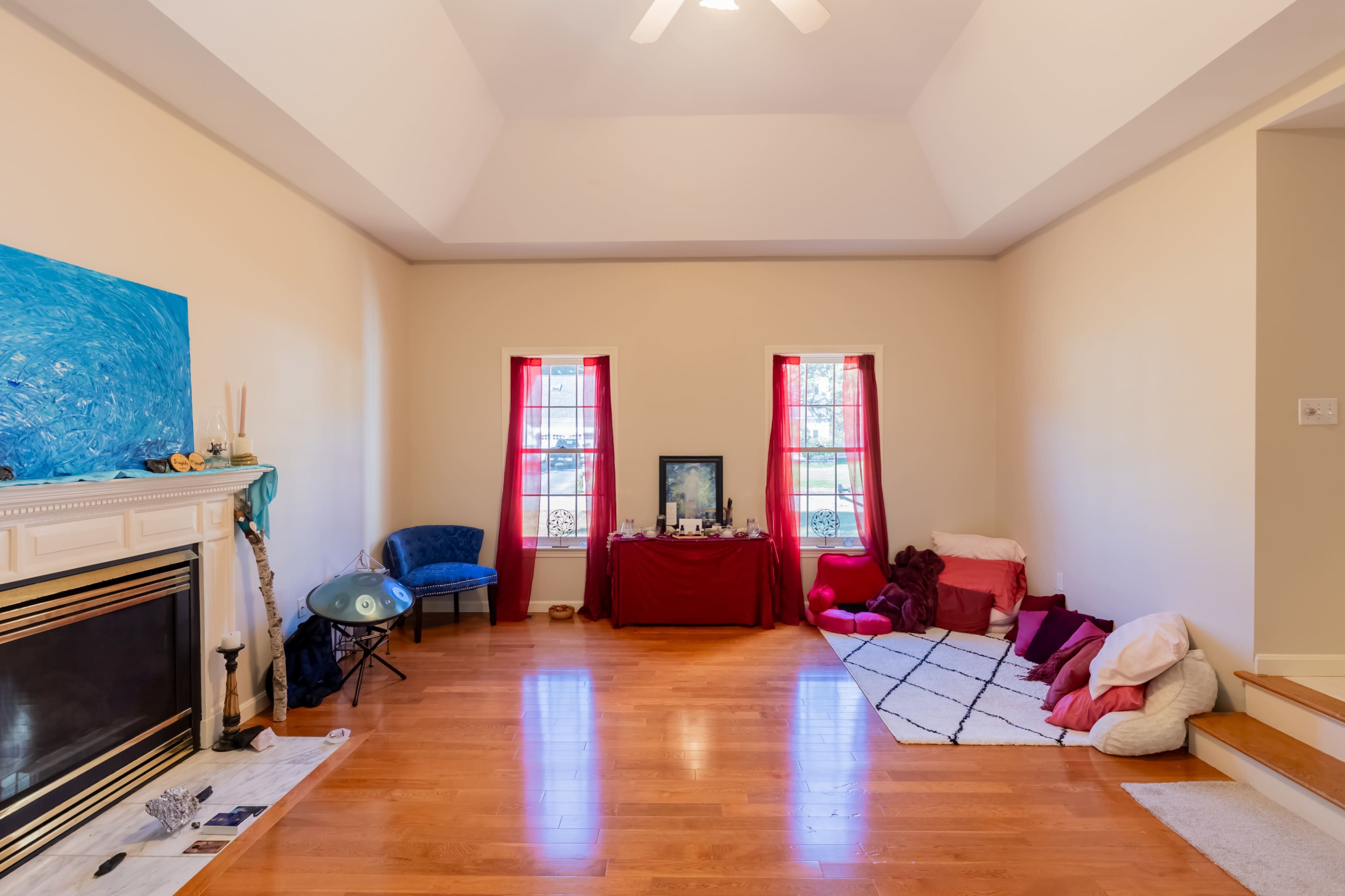 A spacious living room features hardwood floors, two windows with red curtains, and a fireplace with decorative items.