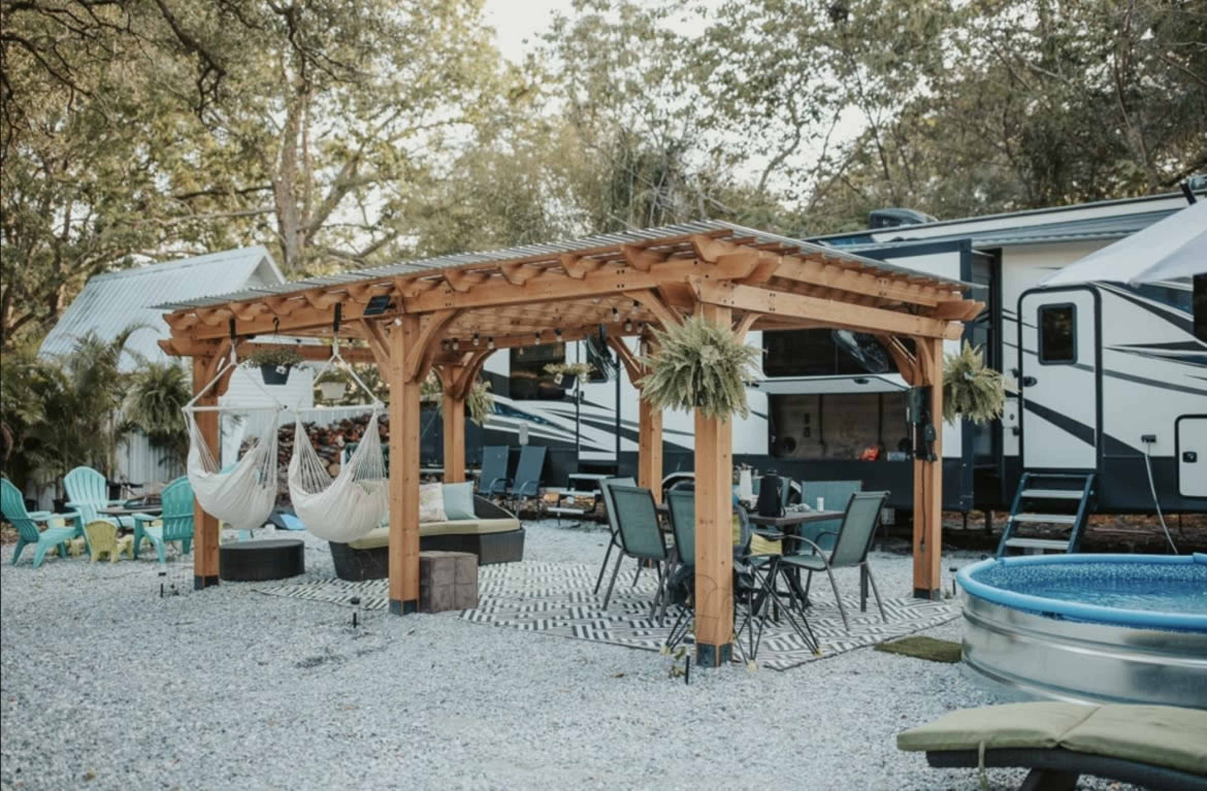 A wooden pergola with hanging hammocks is set in a gravel area alongside a recreational vehicle, surrounded by outdoor furniture and a small pool.