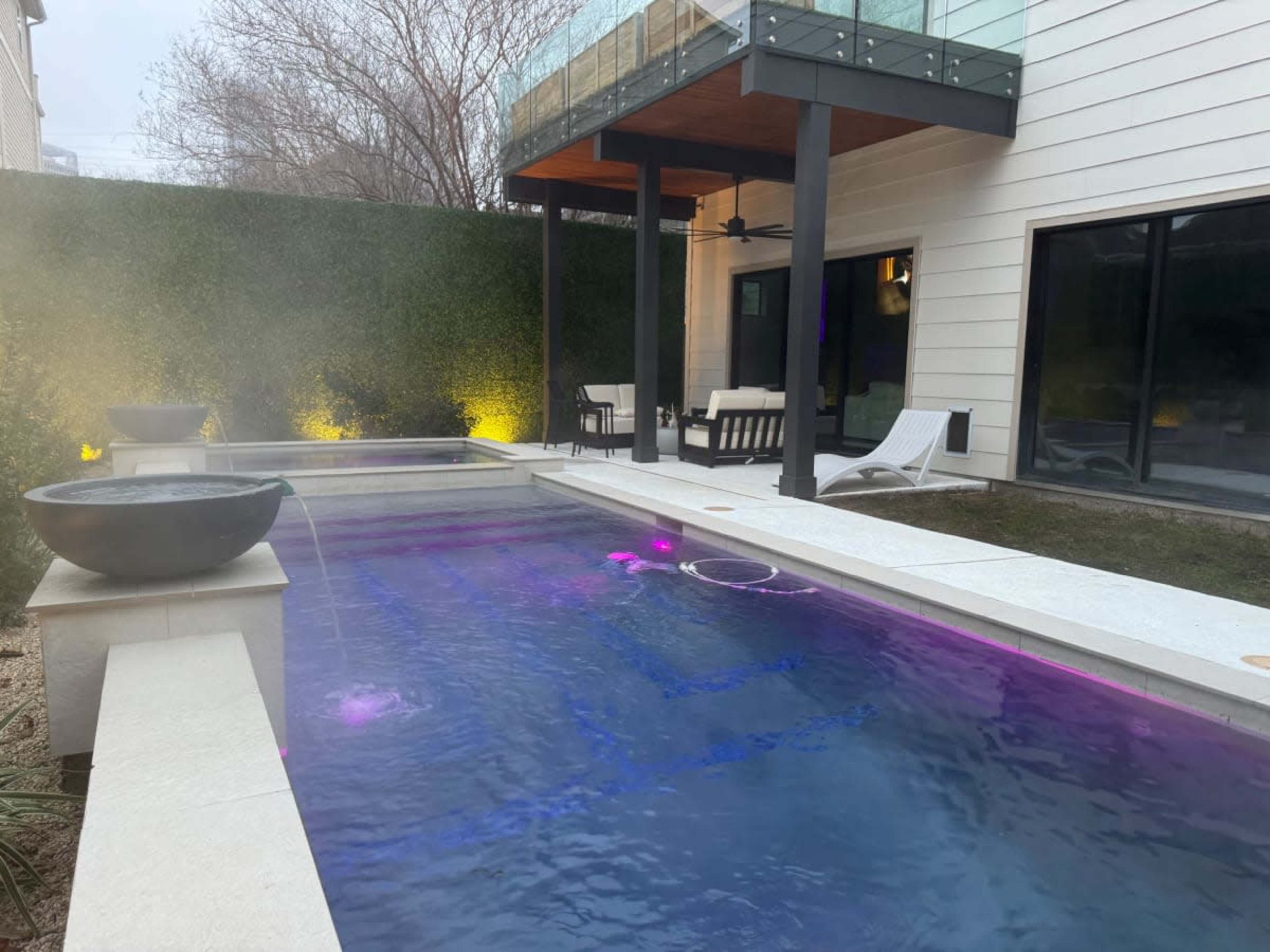 The image shows a modern backyard with a swimming pool featuring illuminated water and a lounge area under a covered patio.