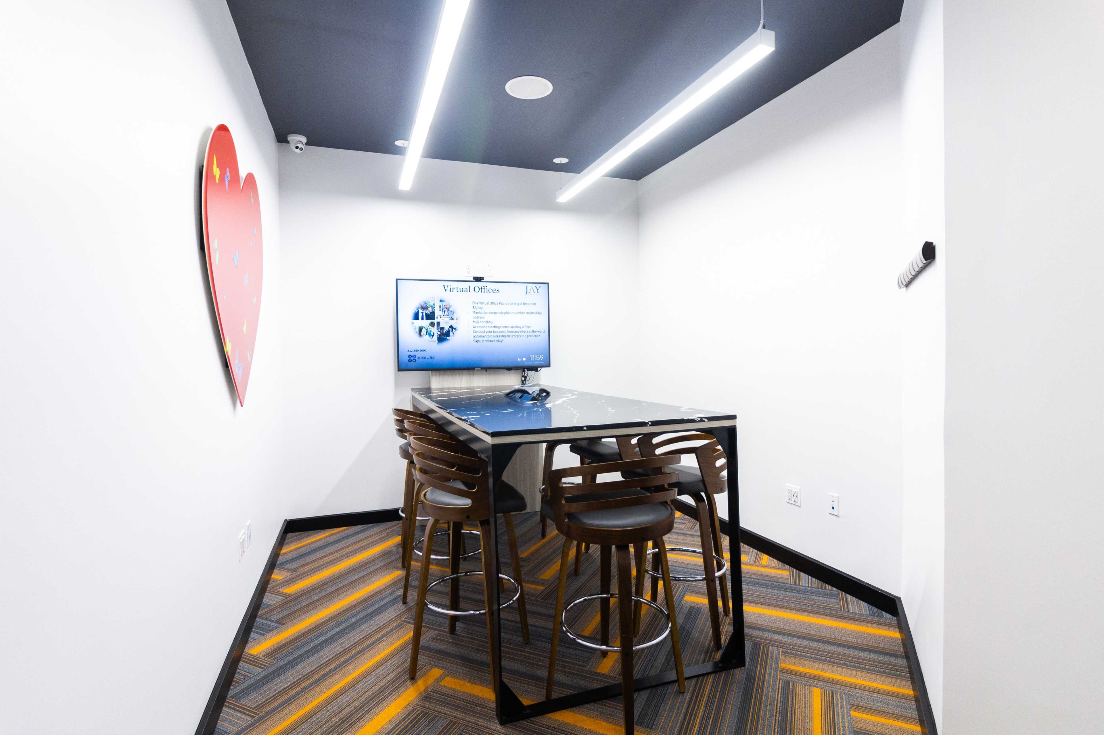 A modern meeting room features a glass table surrounded by high stools, a television screen displaying a presentation, and a heart-shaped decoration on the wall.