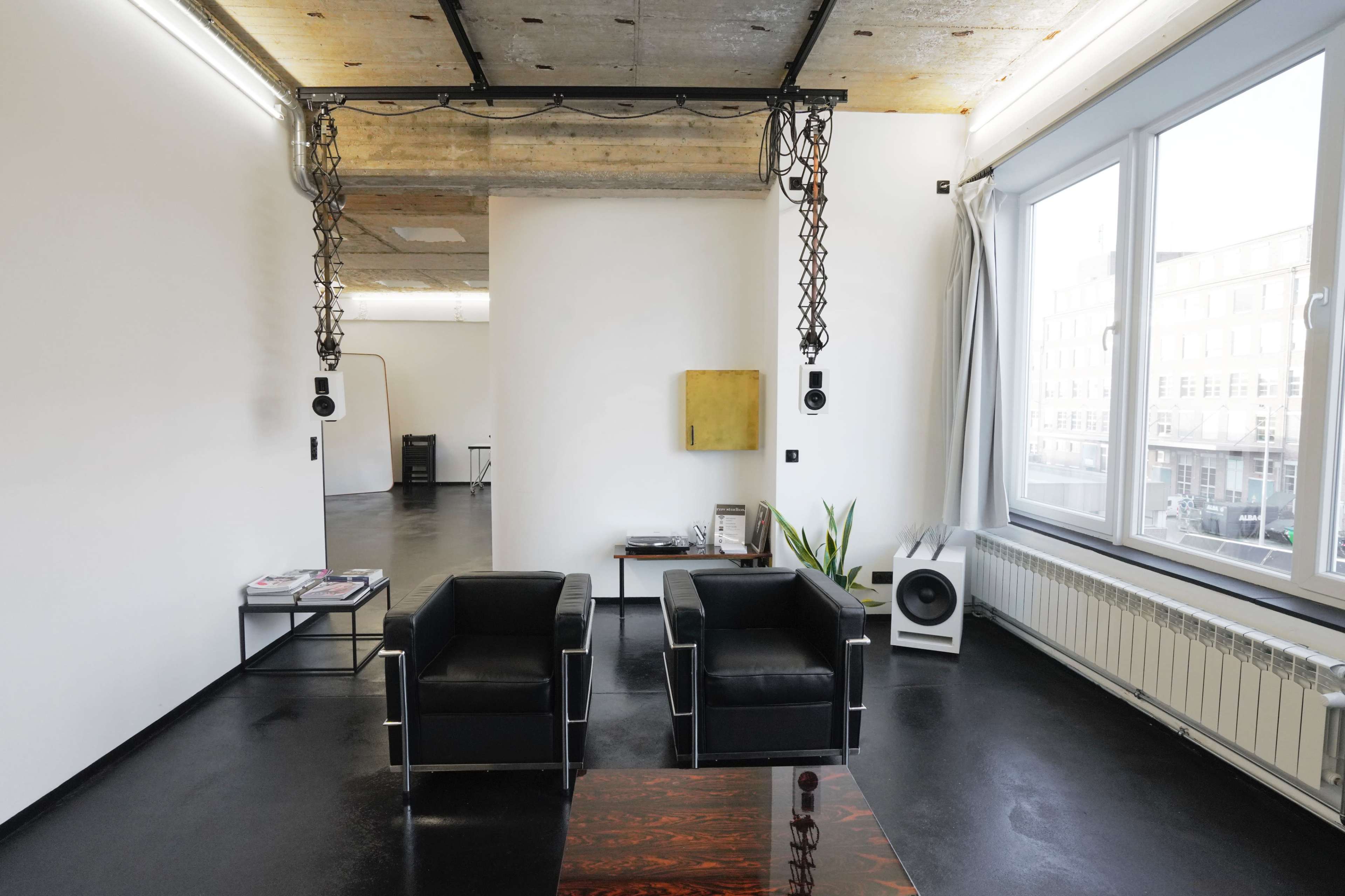 A modern, minimalist interior space featuring two black armchairs facing a glass coffee table, with large windows and industrial-style exposed elements.