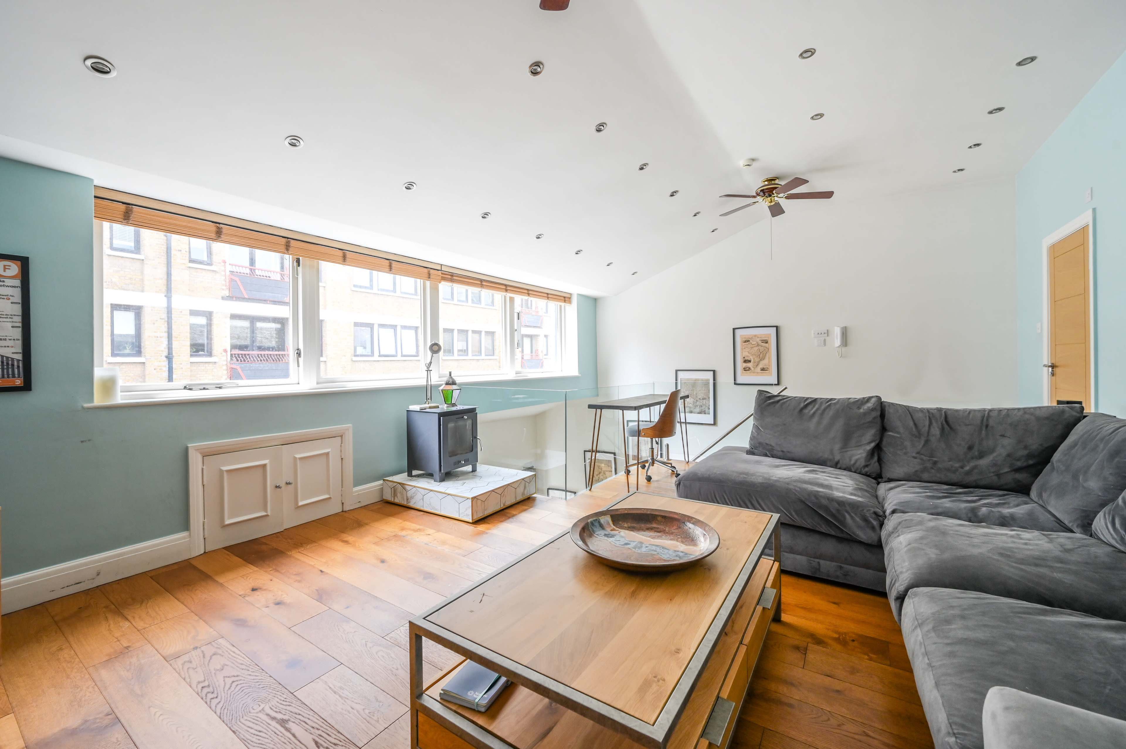 The image shows a bright, modern living room with a gray sectional sofa, wooden flooring, a small dining setup, and large windows allowing natural light to enter.