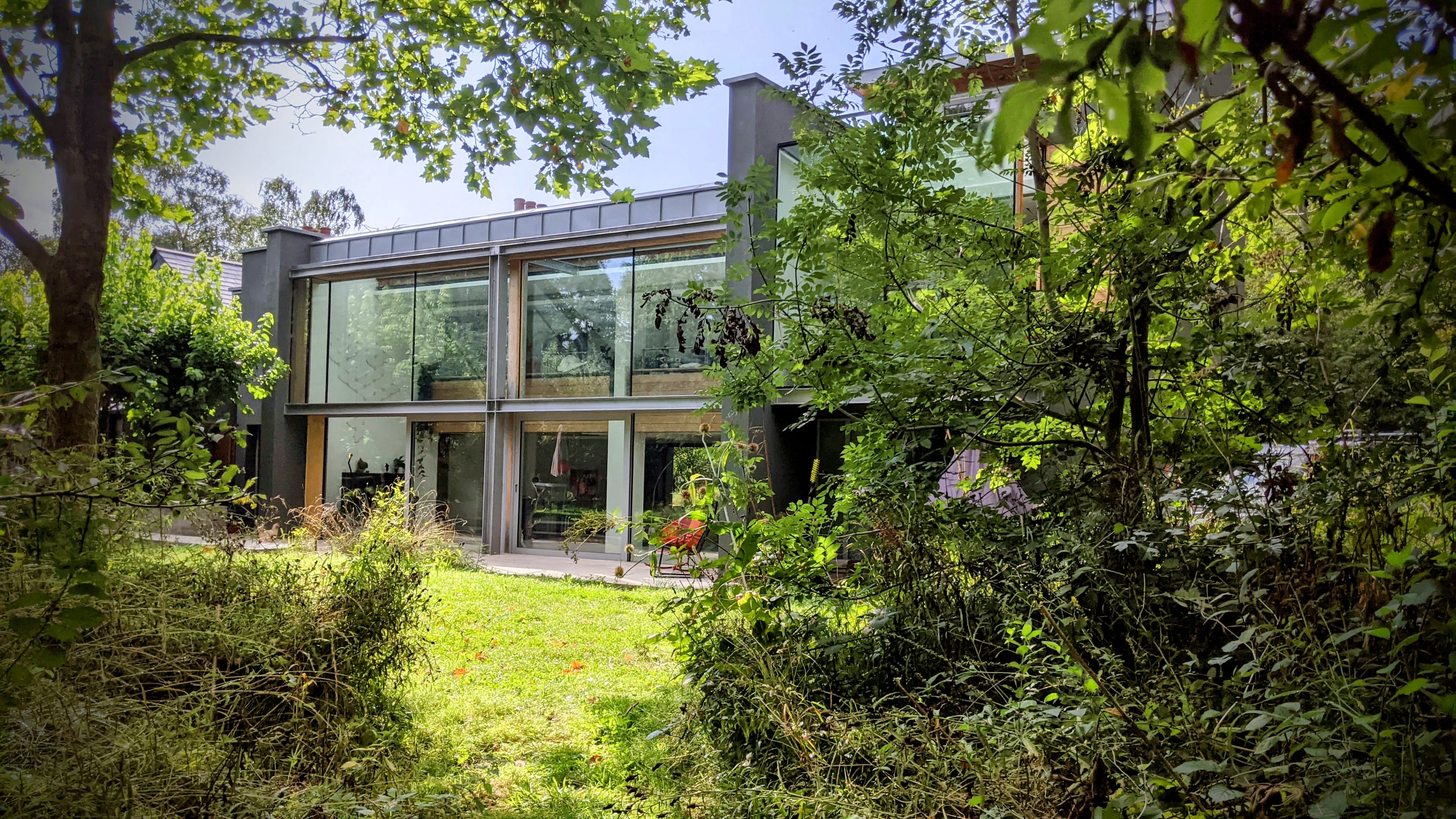 A modern house with large glass windows is surrounded by trees and greenery.