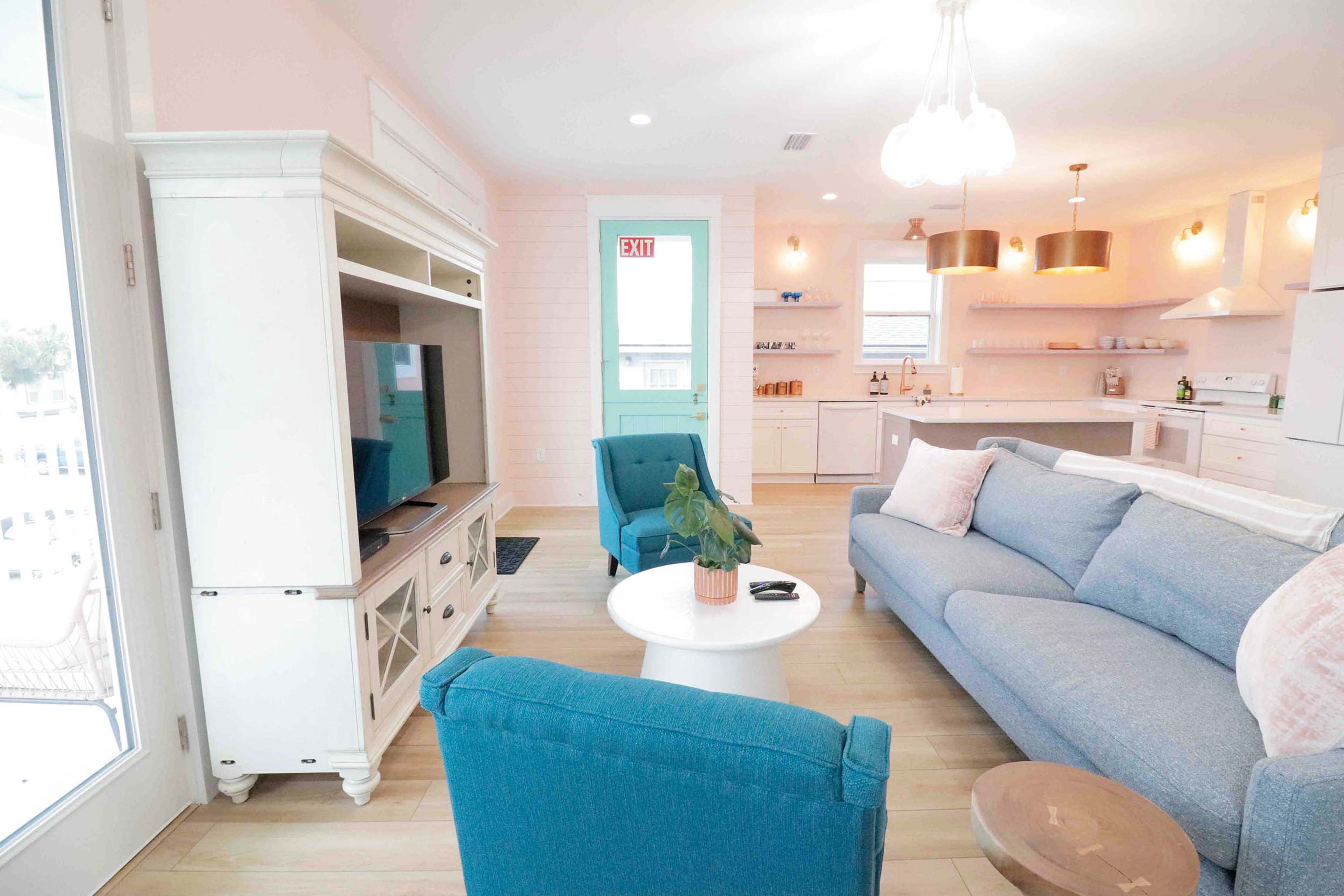 A brightly lit living room with a blue couch, turquoise chairs, a white coffee table, and a modern kitchen in the background.