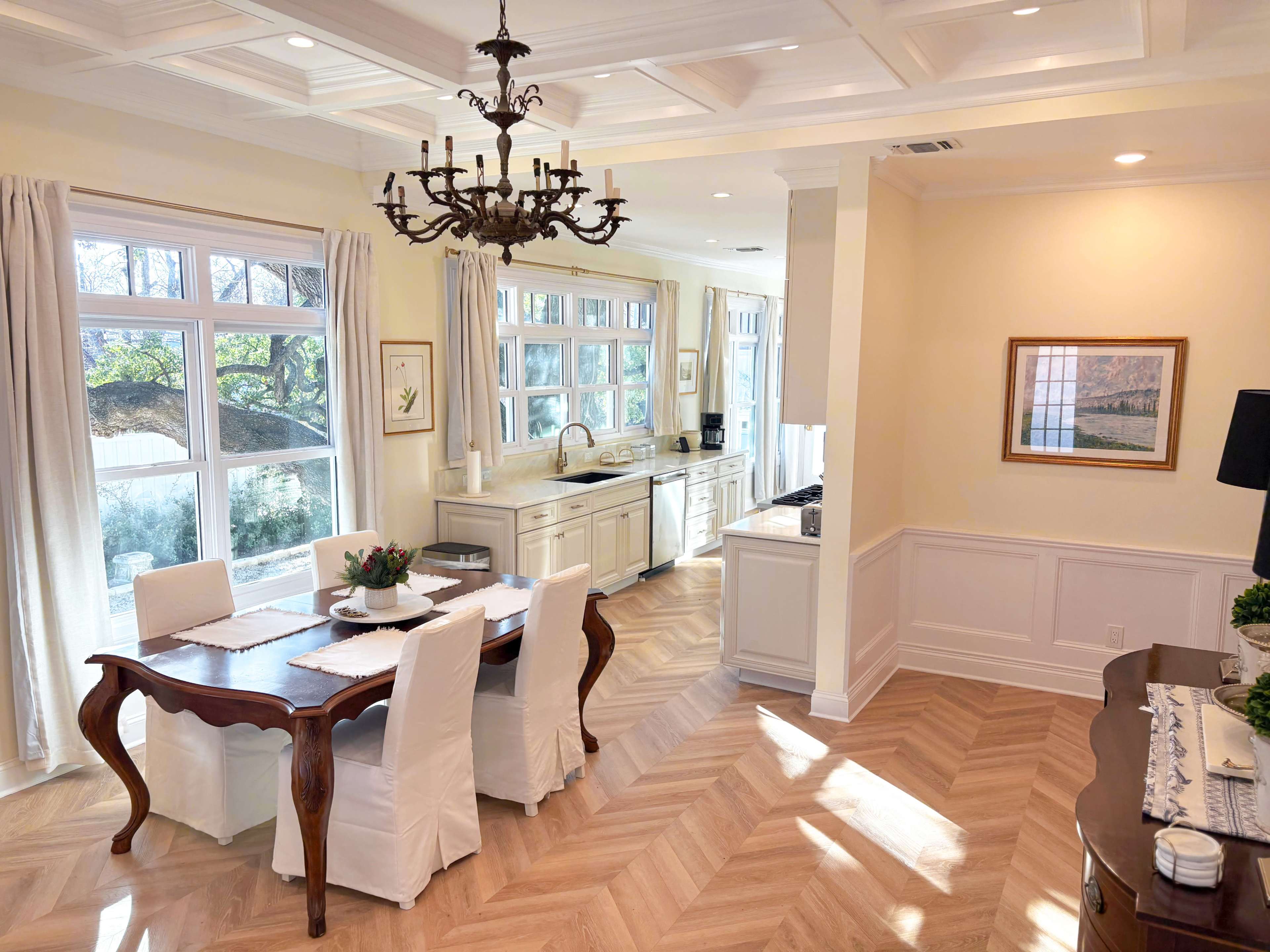 The image depicts a bright, airy kitchen and dining area featuring a wooden dining table with white chairs, large windows allowing natural light, and classic cabinetry.