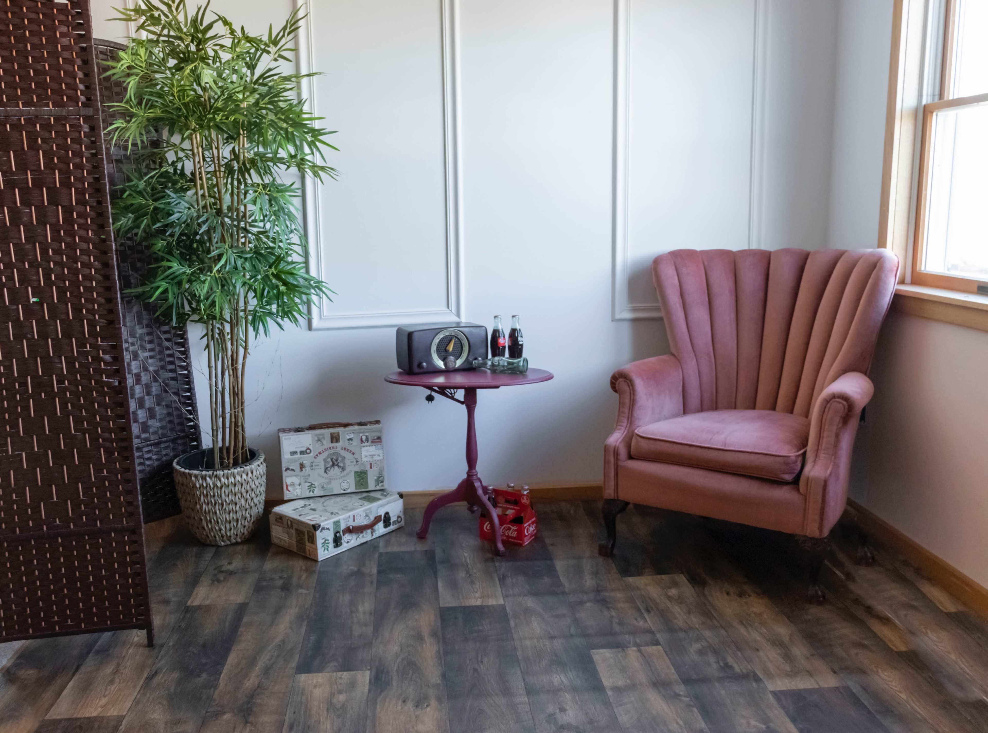A corner of a room with a pink armchair, a small round table holding two bottles, a potted plant, and a patterned box on the wooden floor.
