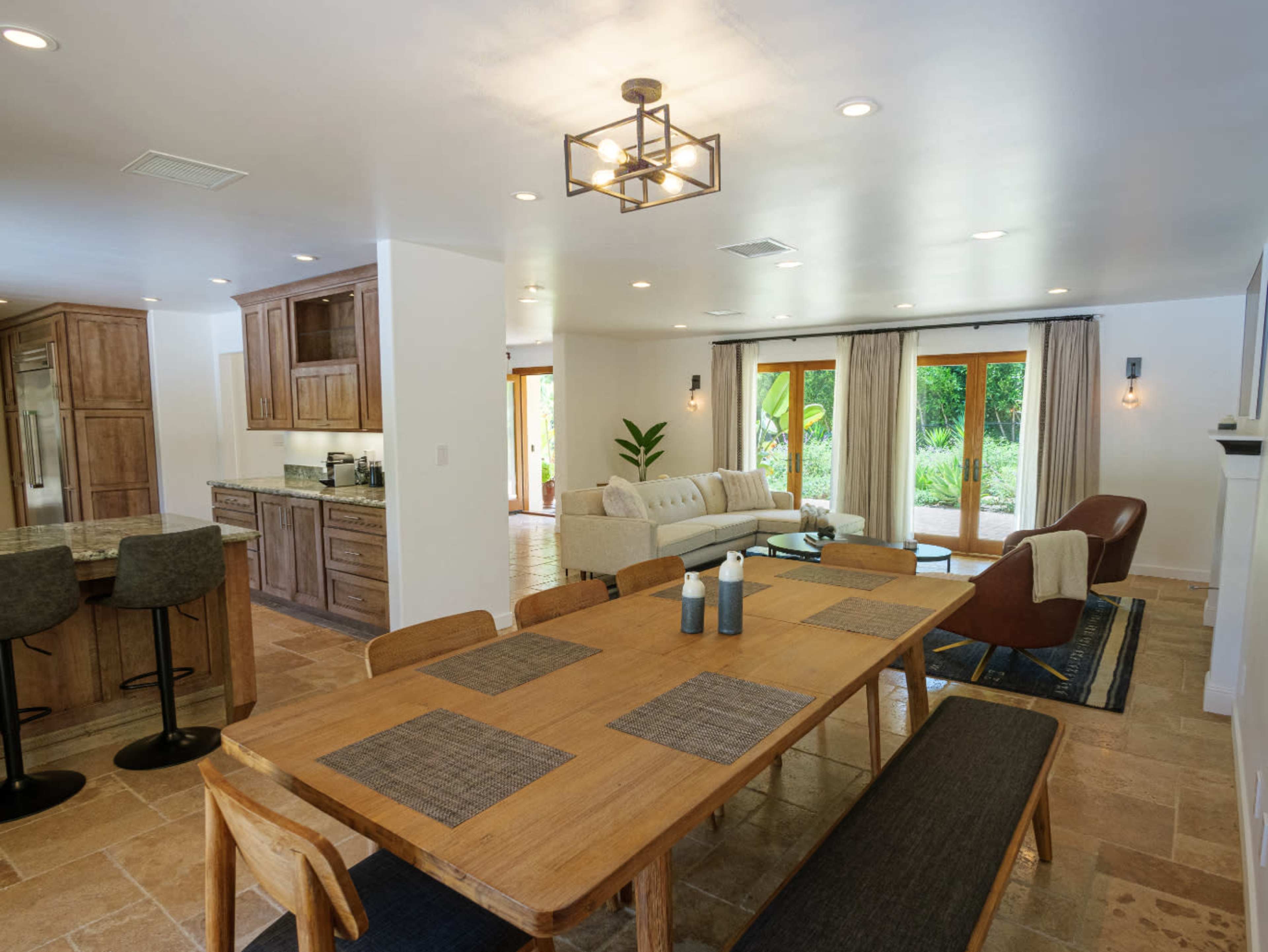 A modern dining area with a wooden table set for six, adjacent to a kitchen and a comfortable living space featuring plush seating and large windows.