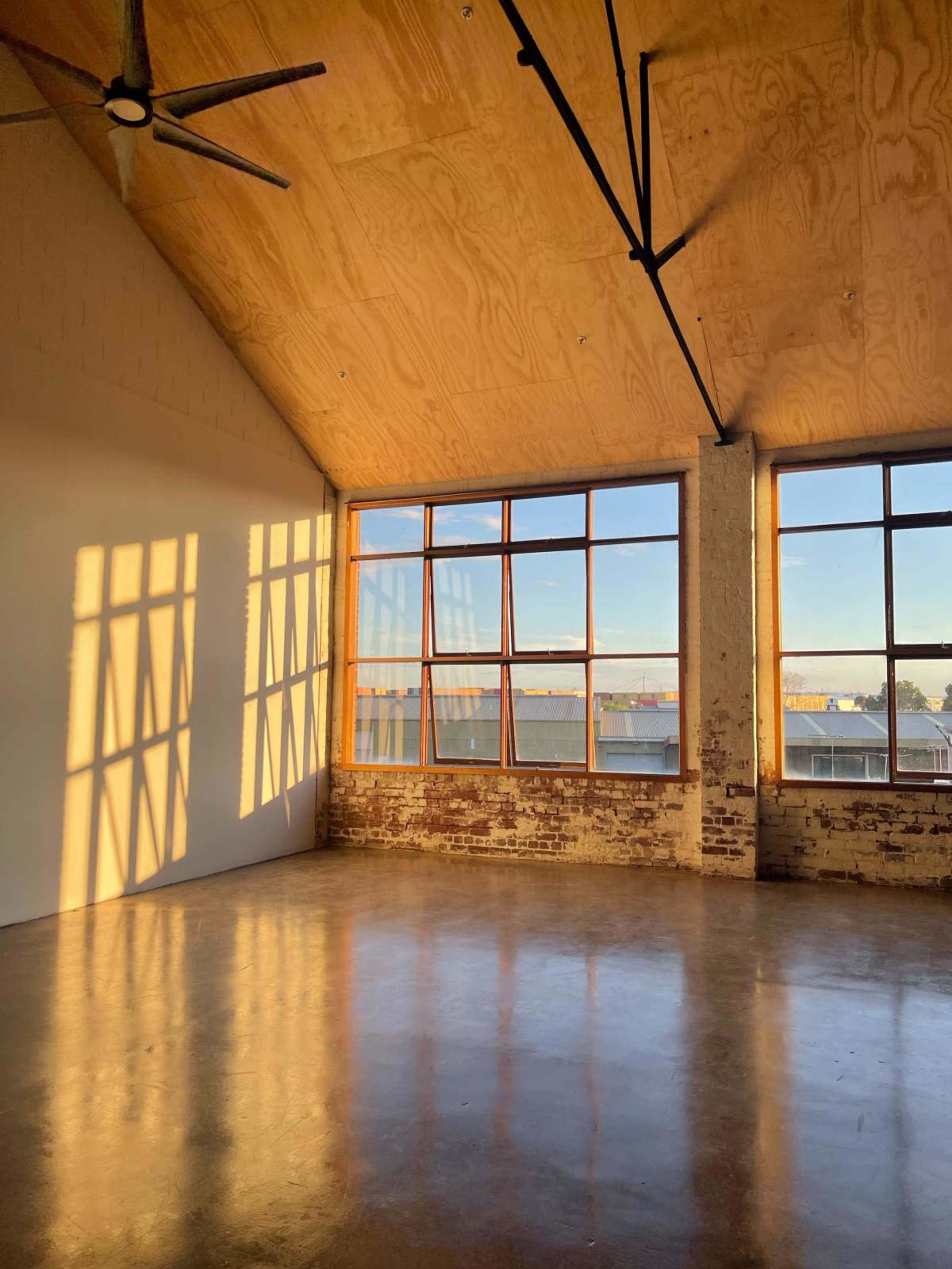 The image shows a spacious room with large windows, wooden ceiling, and sunlight casting shadows on the polished floor.