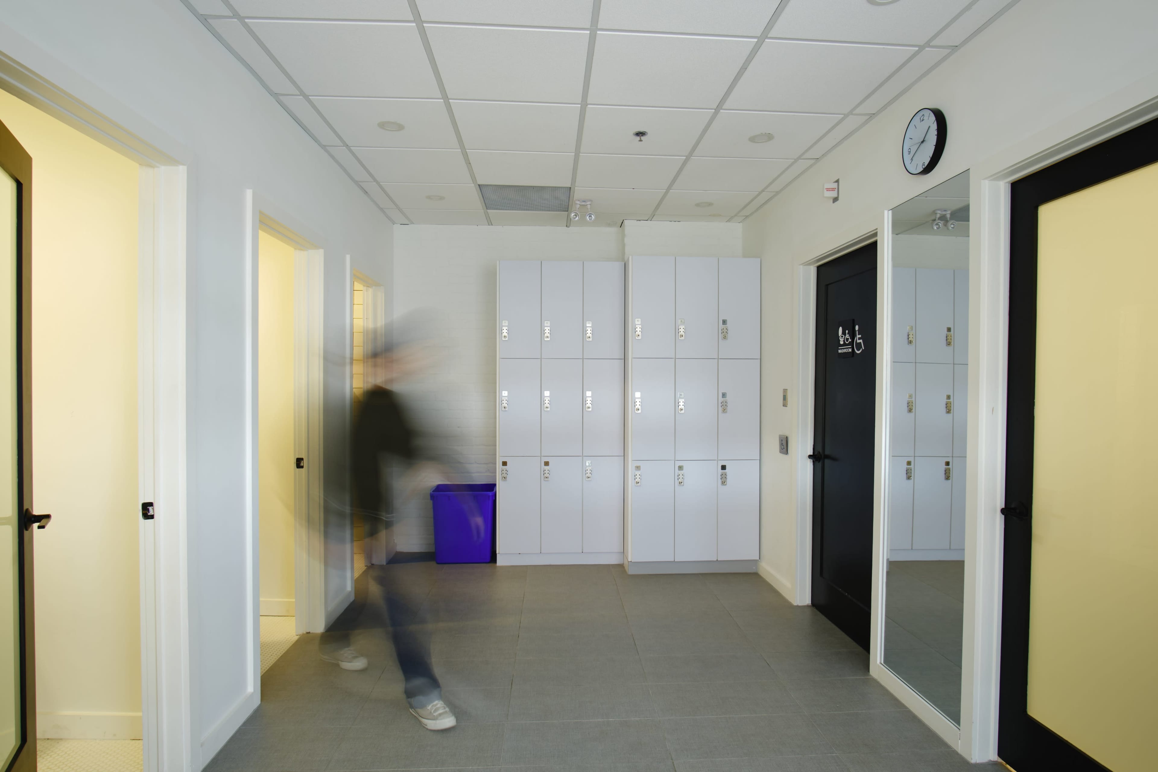 A hallway features several white lockers, doors to rooms on either side, and a clock on the wall, with a person walking quickly in the background.