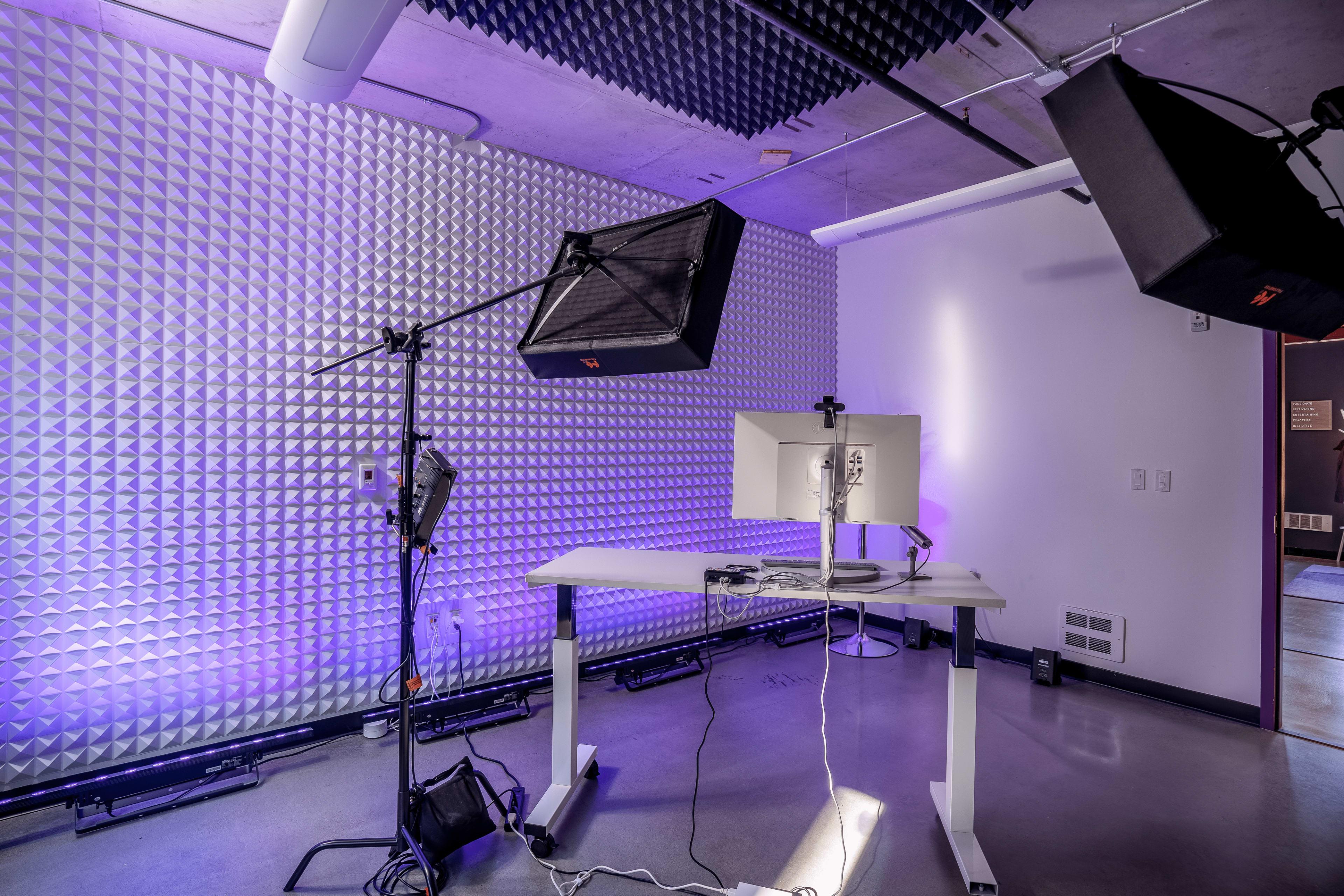 A modern workspace featuring a standing desk with a monitor, surrounded by a soundproof wall and studio lighting equipment.