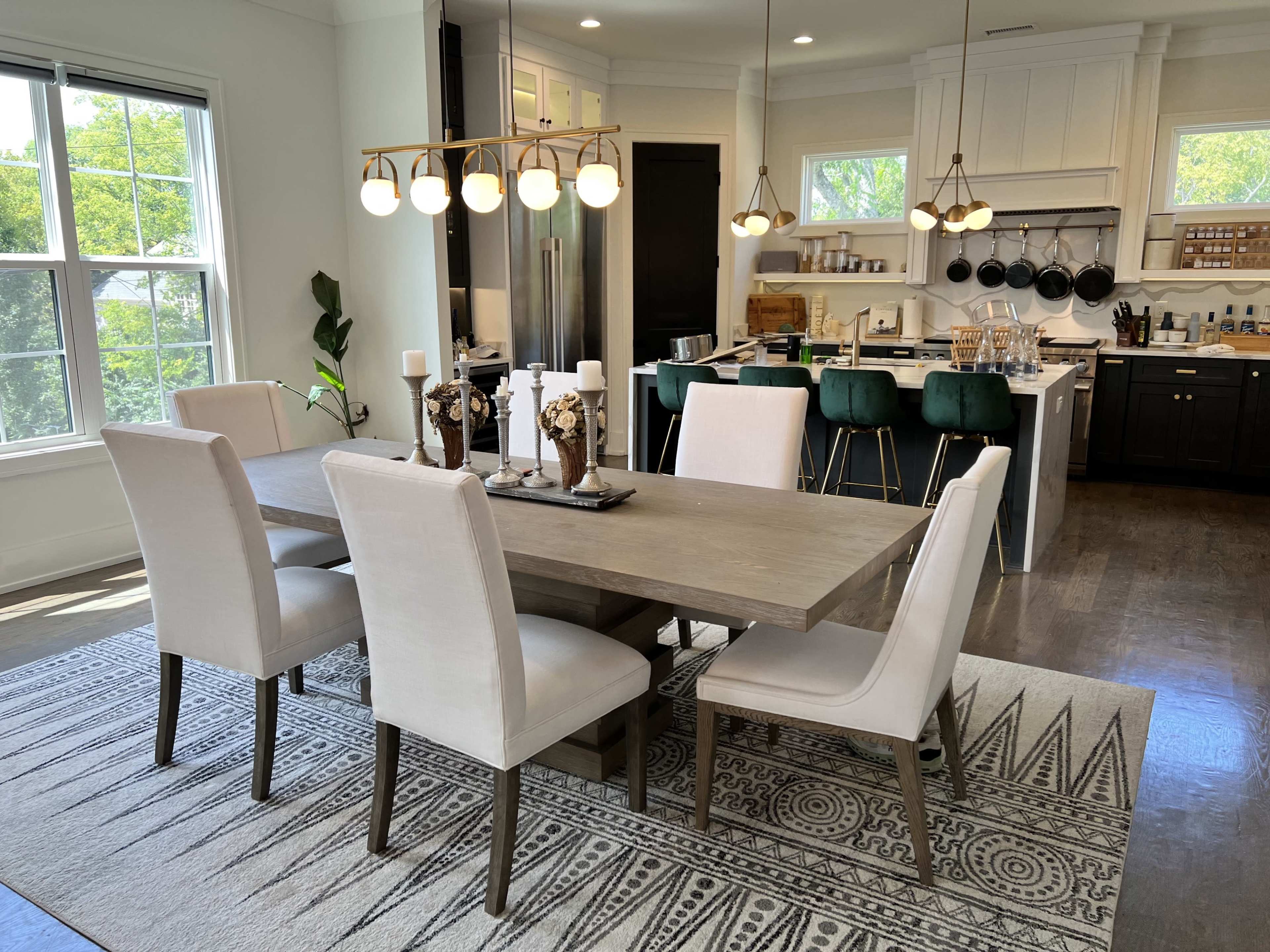 A modern dining area features a wooden table surrounded by upholstered chairs, with a kitchen visible in the background.