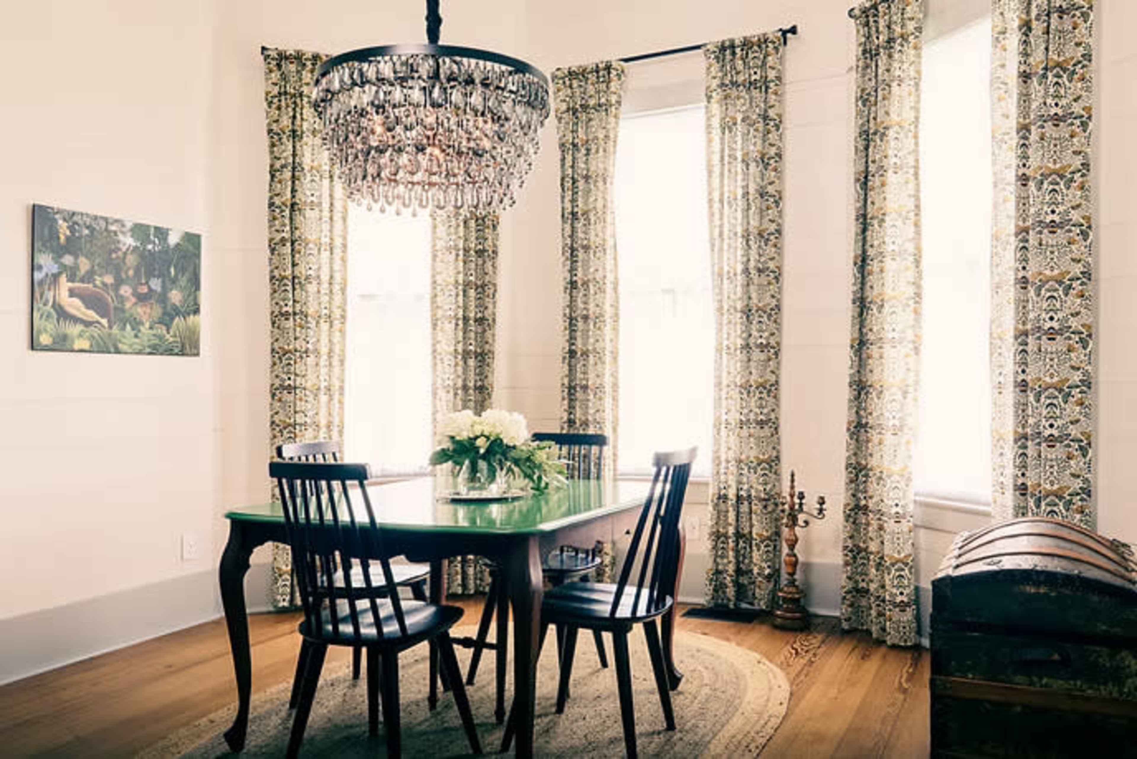 A dining room features a round table surrounded by four black chairs, with a chandelier overhead and large windows dressed in patterned curtains.