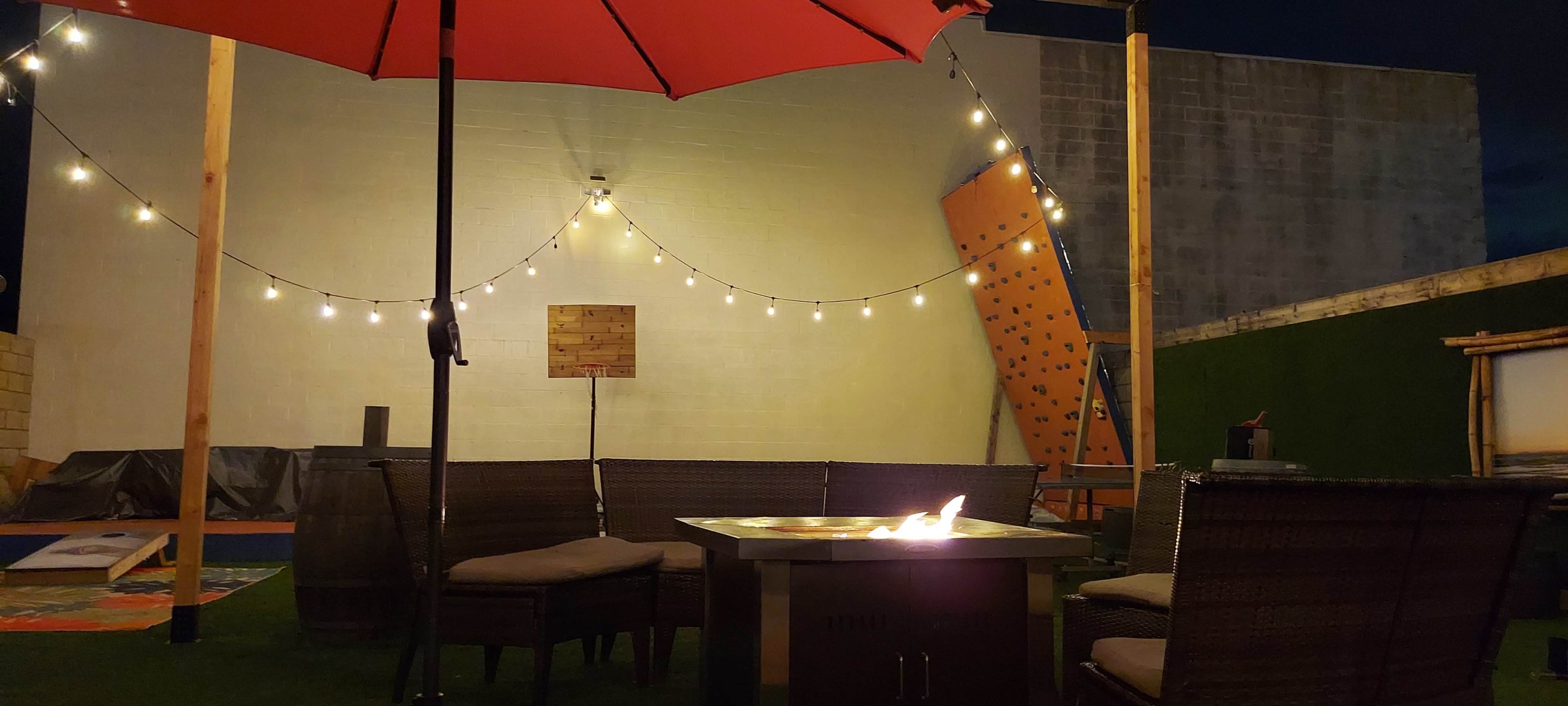 A patio area features a fire pit surrounded by wicker seating, with an umbrella and string lights overhead, in front of a climbing wall.