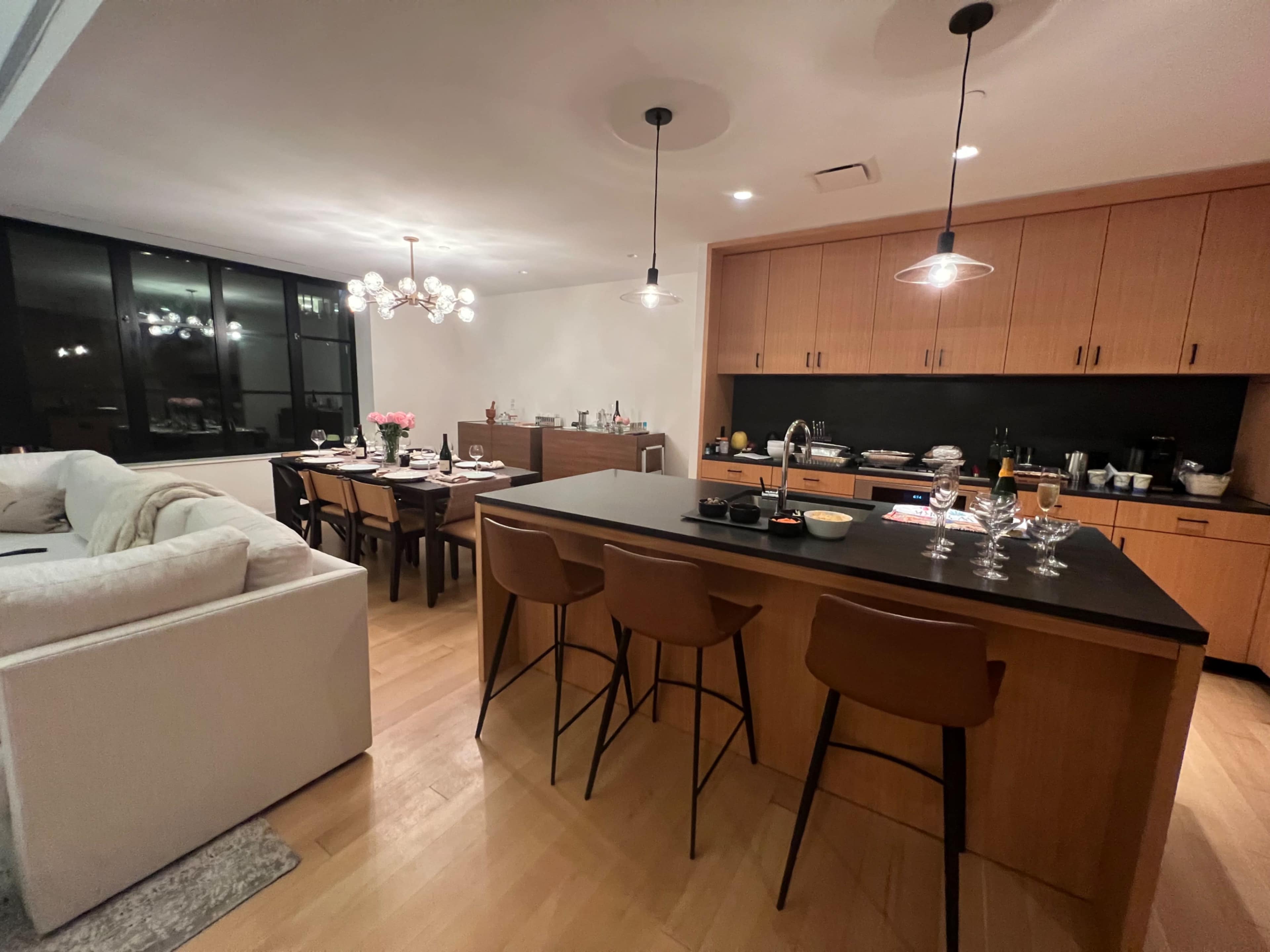 A modern kitchen and dining area features a central island with bar stools, a dining table set for eight, and contemporary lighting fixtures.