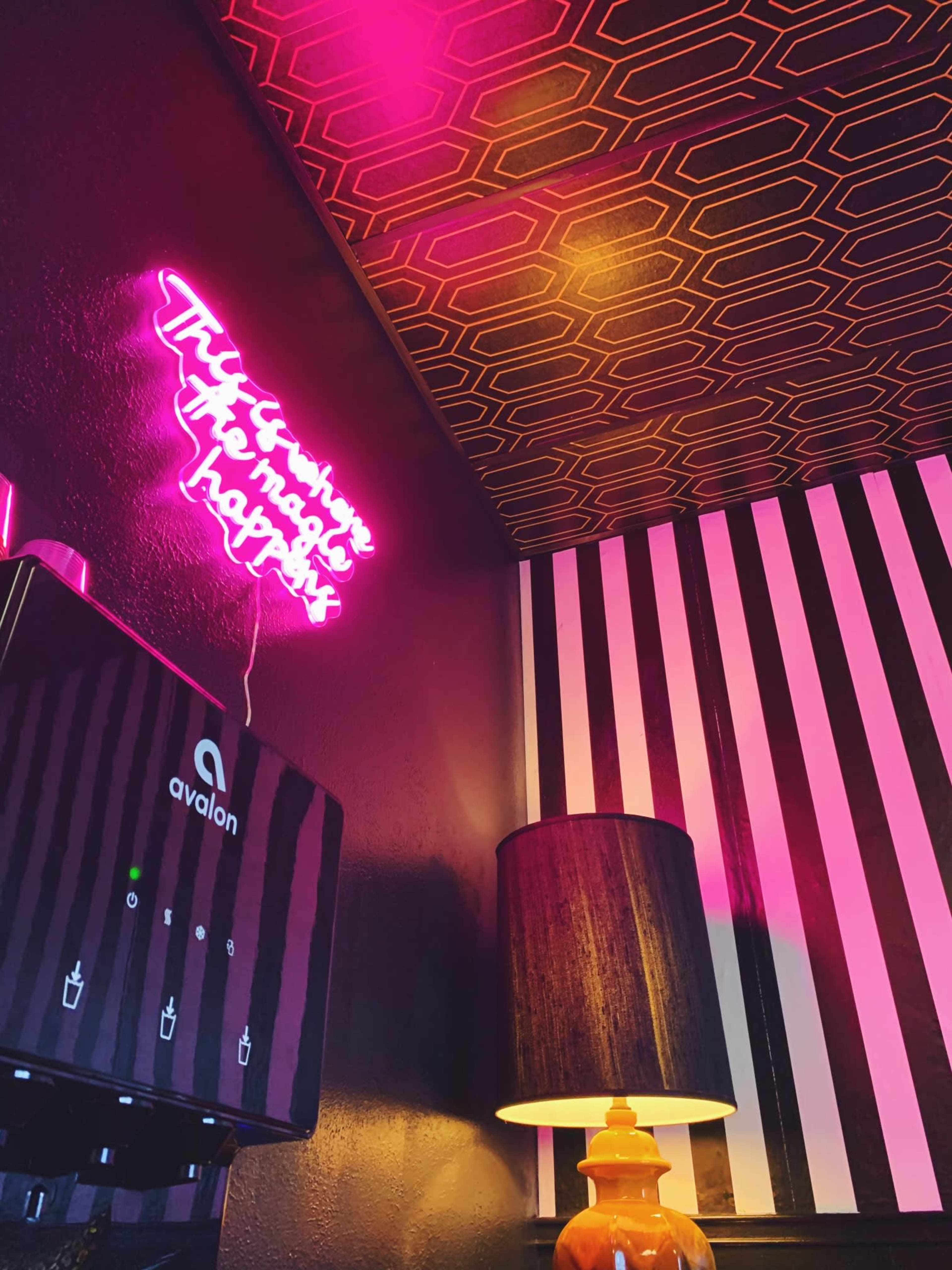A neon sign reading "There's nowhere to find happiness" hangs above a drink dispenser against a patterned wall with black and white stripes.
