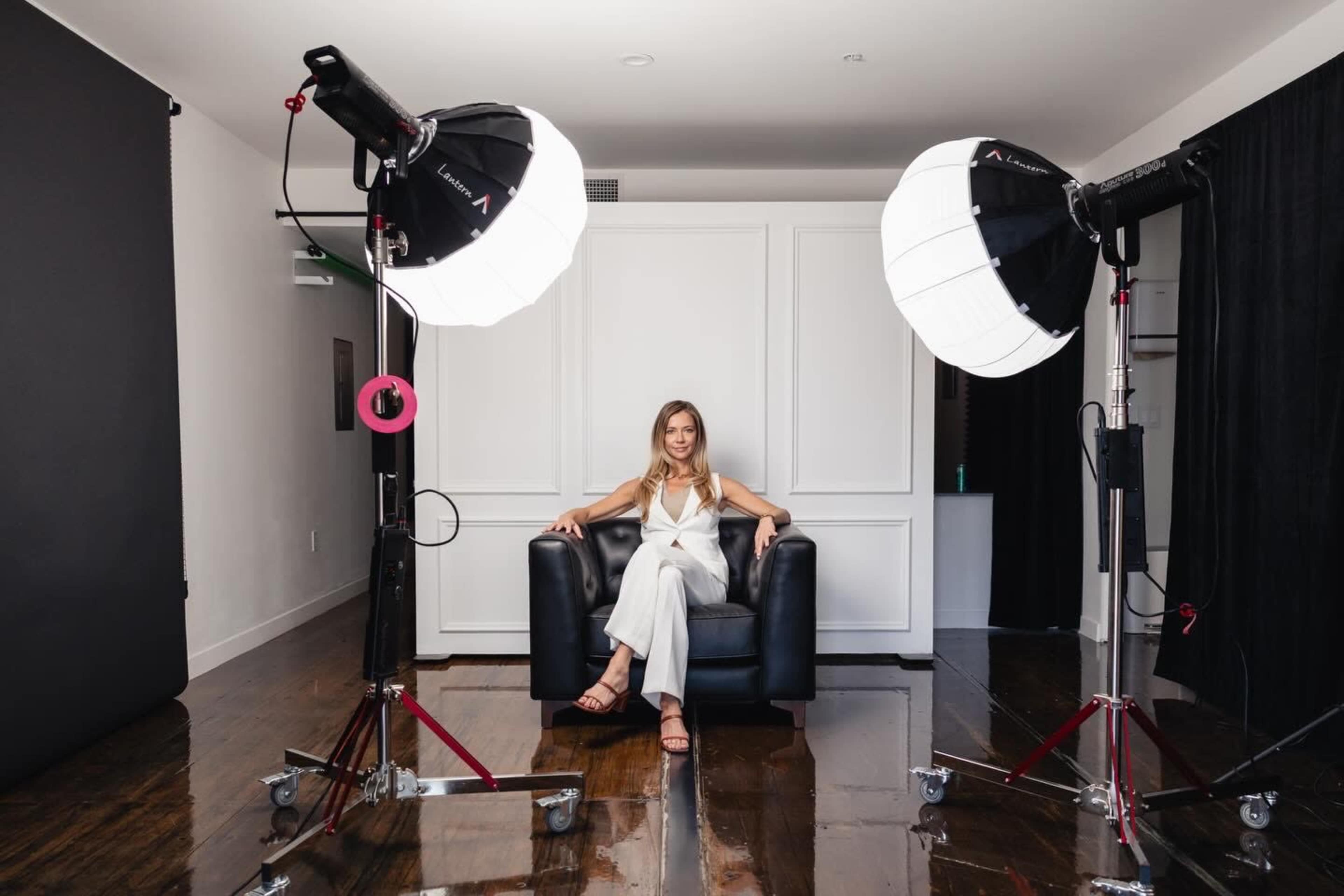 A woman sits in a black chair between two large studio lights in a photography setting.
