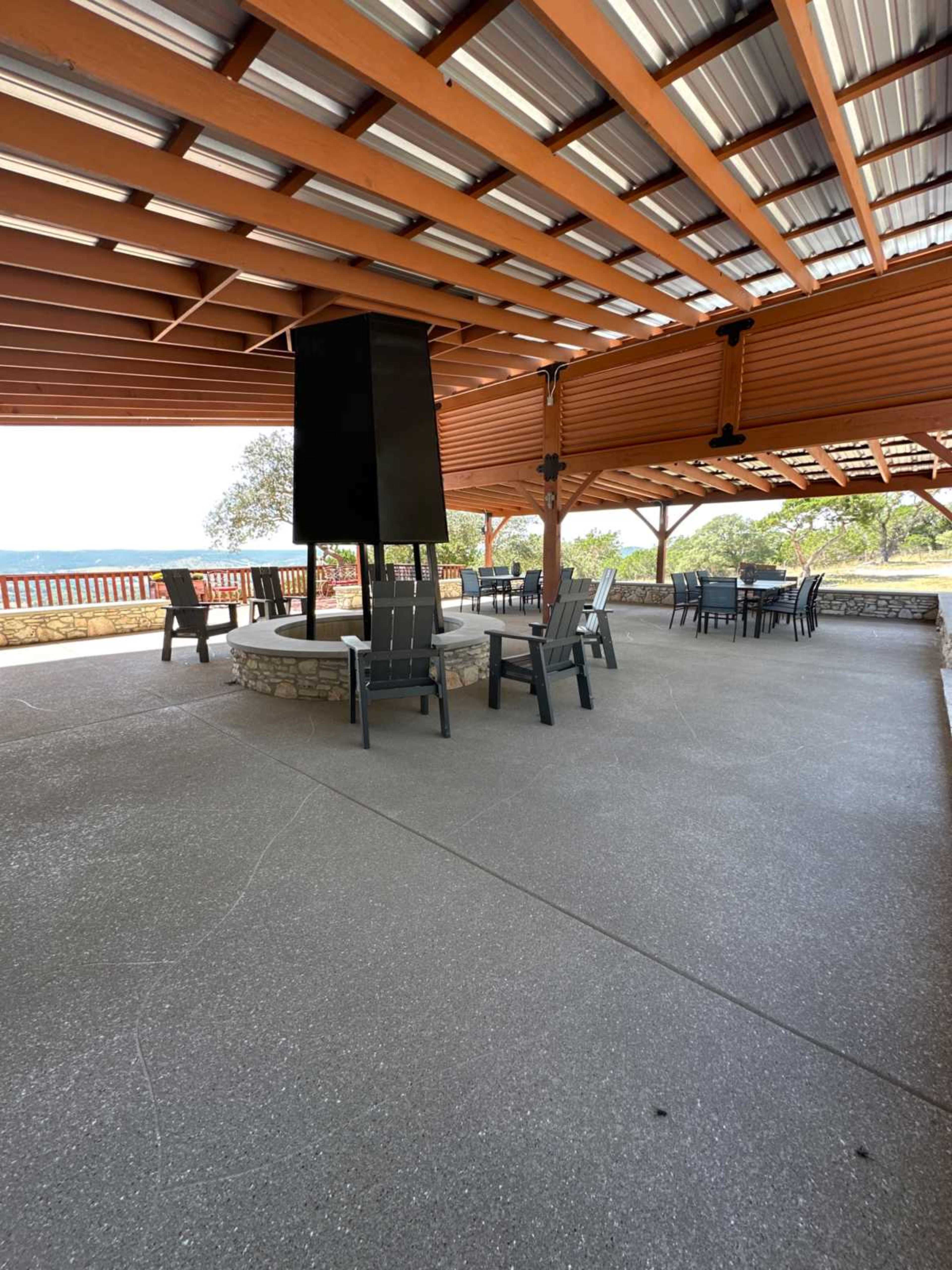 The image shows a shaded outdoor seating area with a circular stone fire pit and metal chairs arranged around it.