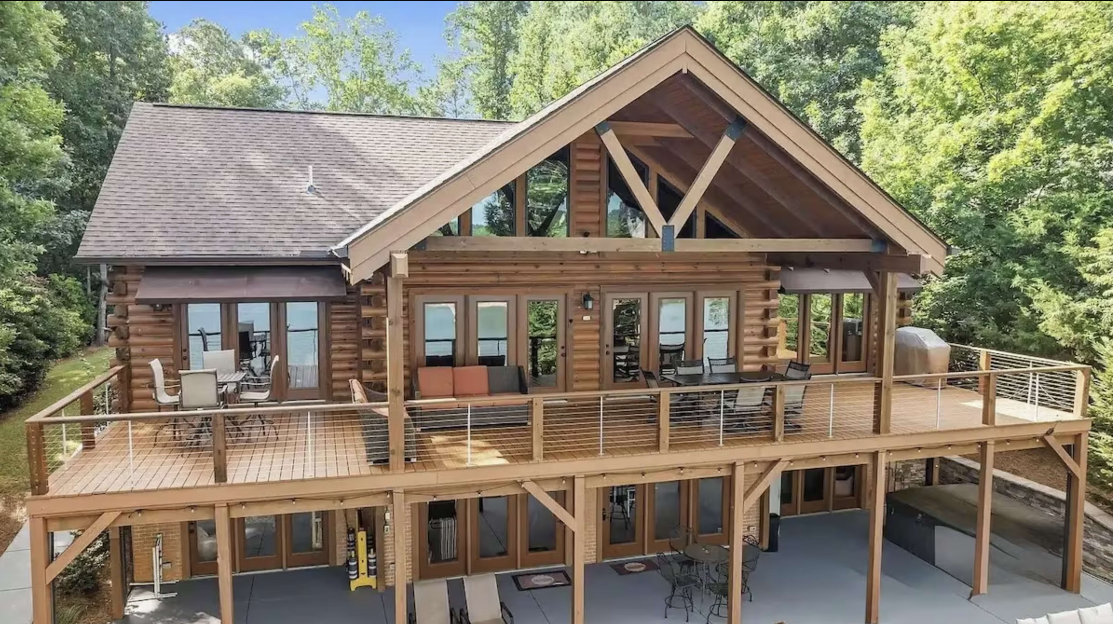 The image shows a large wooden cabin with a prominent deck, featuring multiple windows and a rustic architectural style surrounded by greenery.