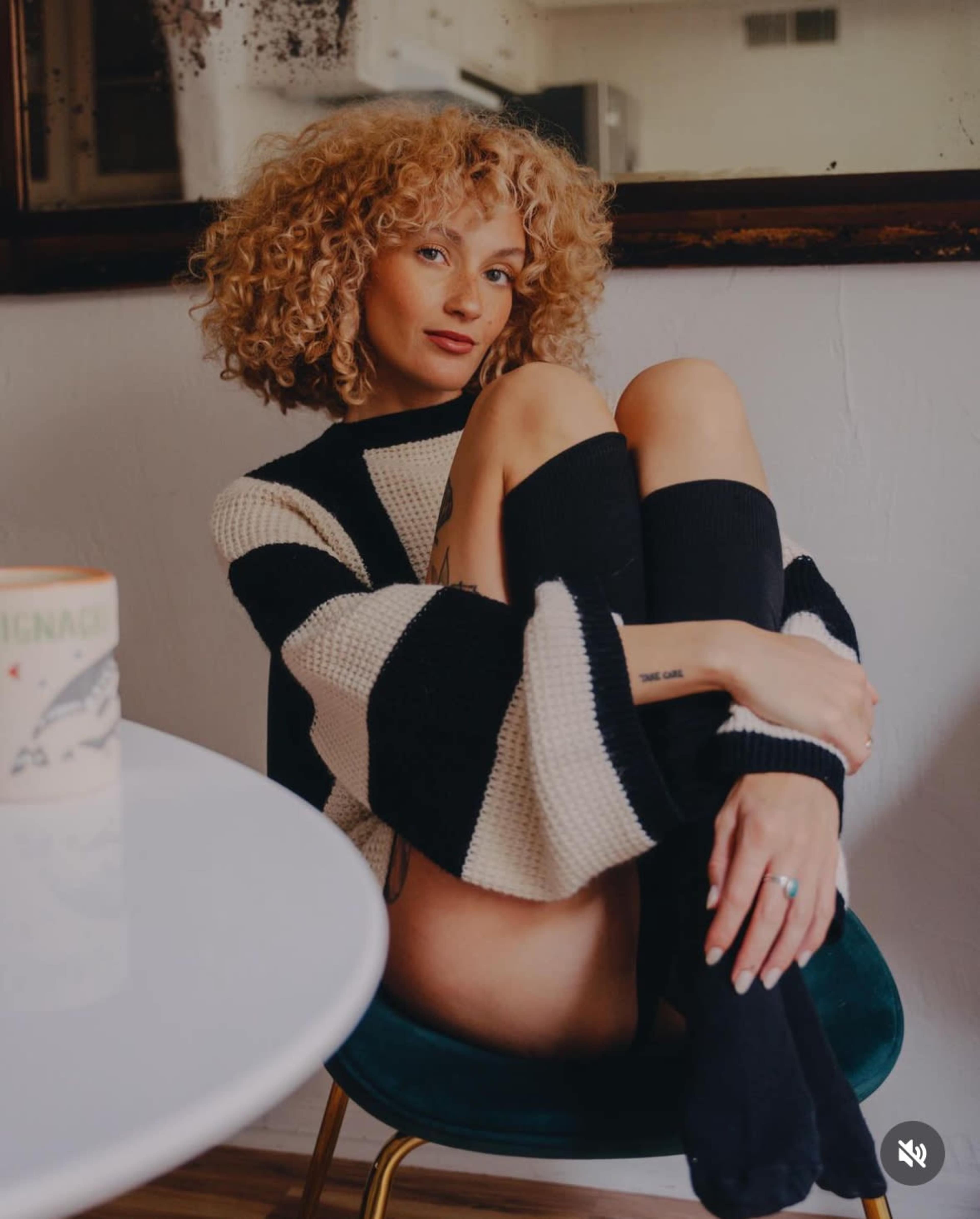 A person with curly hair sits on a chair, hugging their knees with one arm while wearing a striped sweater and knee-high socks.