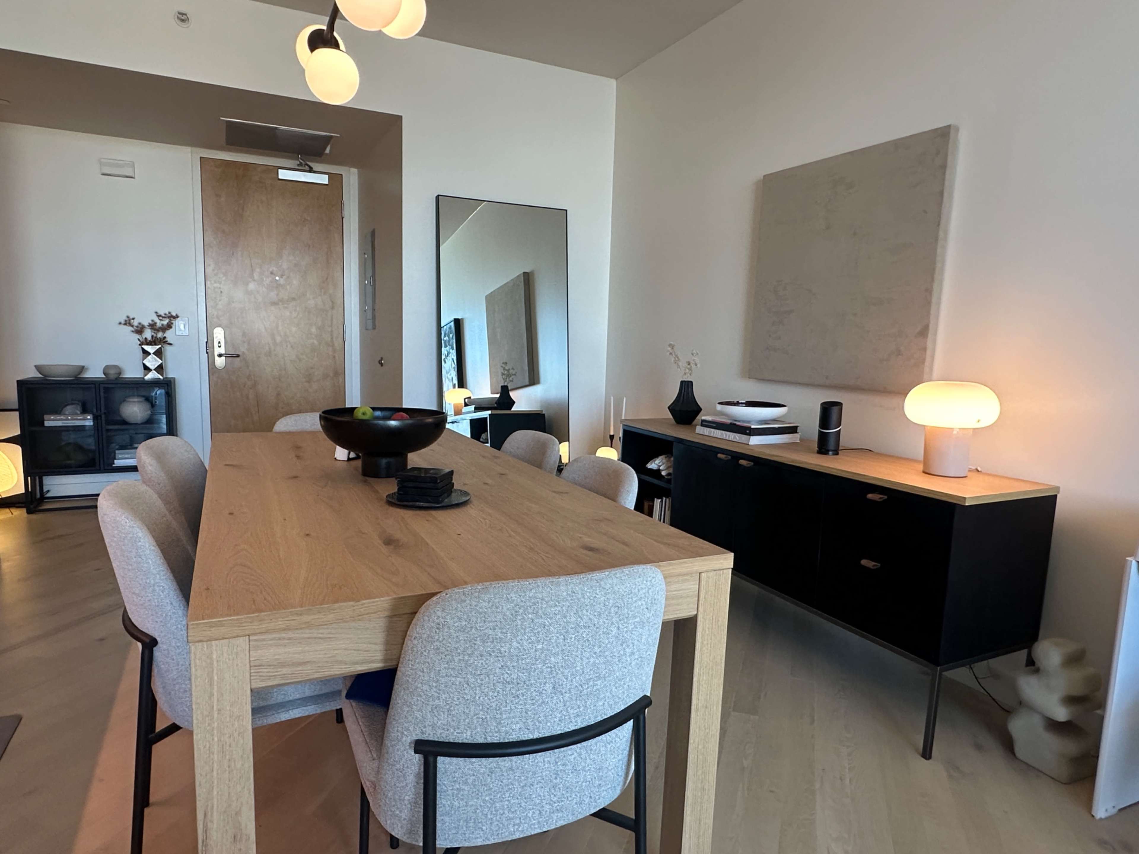 A dining area features a wooden table surrounded by upholstered chairs, with a console, mirror, and wall art in a modern interior.