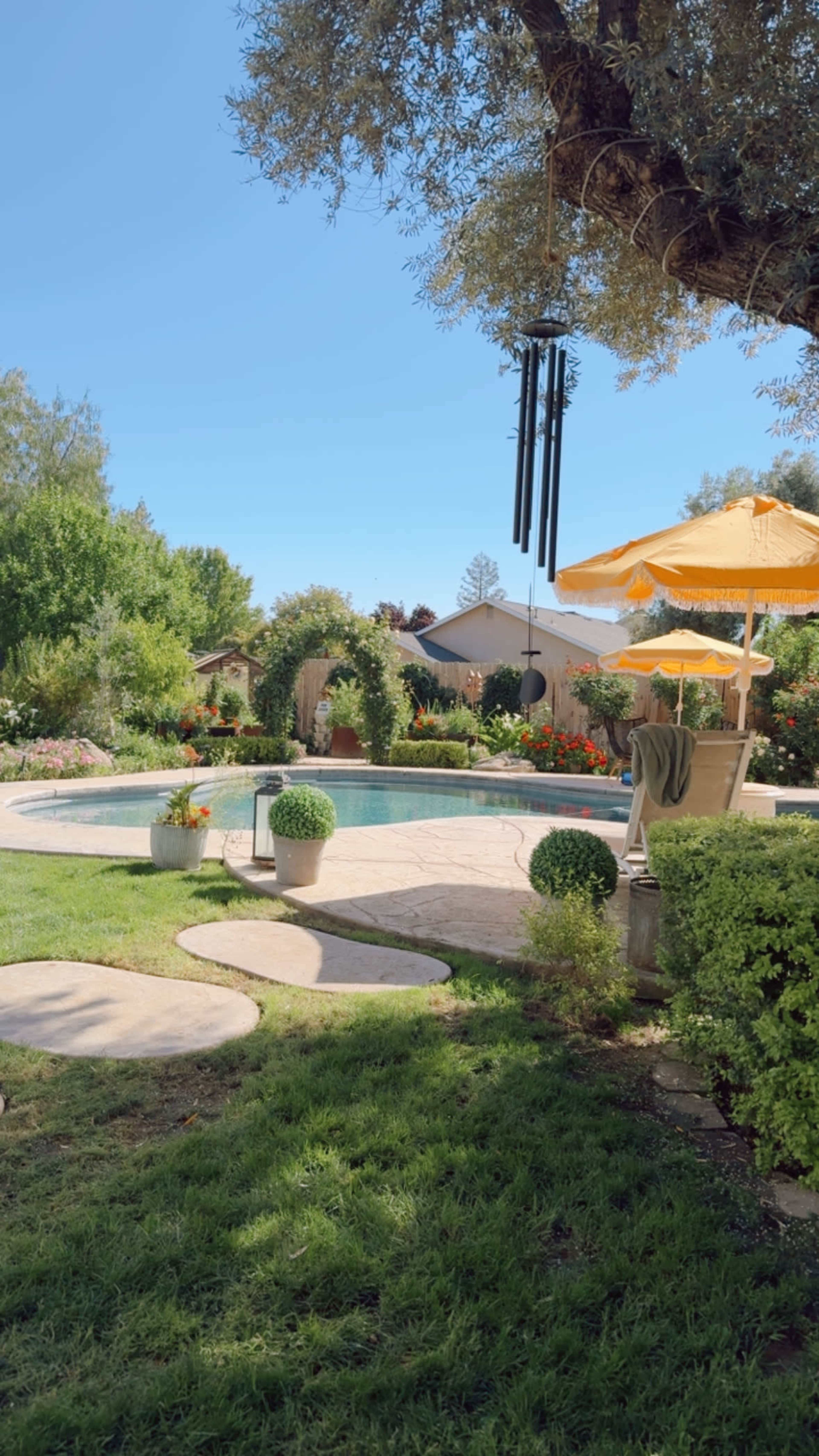 The scene features a beautifully landscaped backyard with a swimming pool, orange umbrellas, and greenery surrounding a stone path.