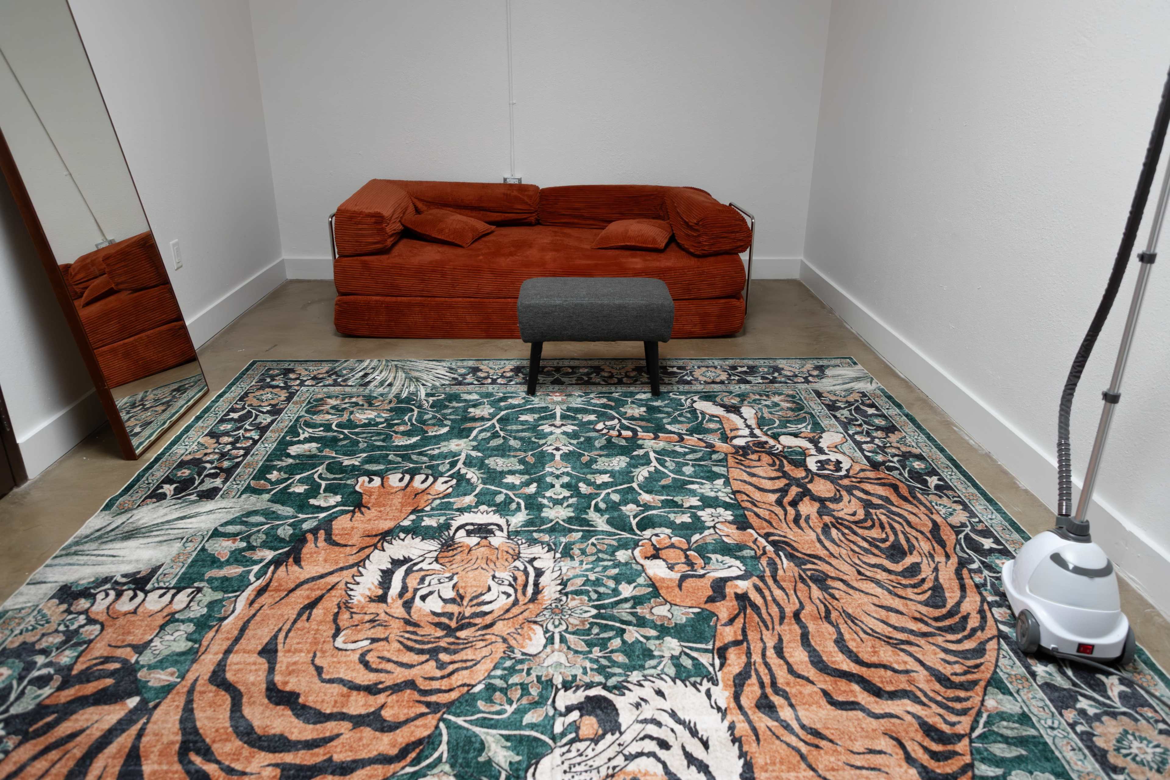 The image shows a sparsely furnished room featuring an orange couch, a small black stool, a large tiger-patterned rug, and a steam cleaner in the corner.