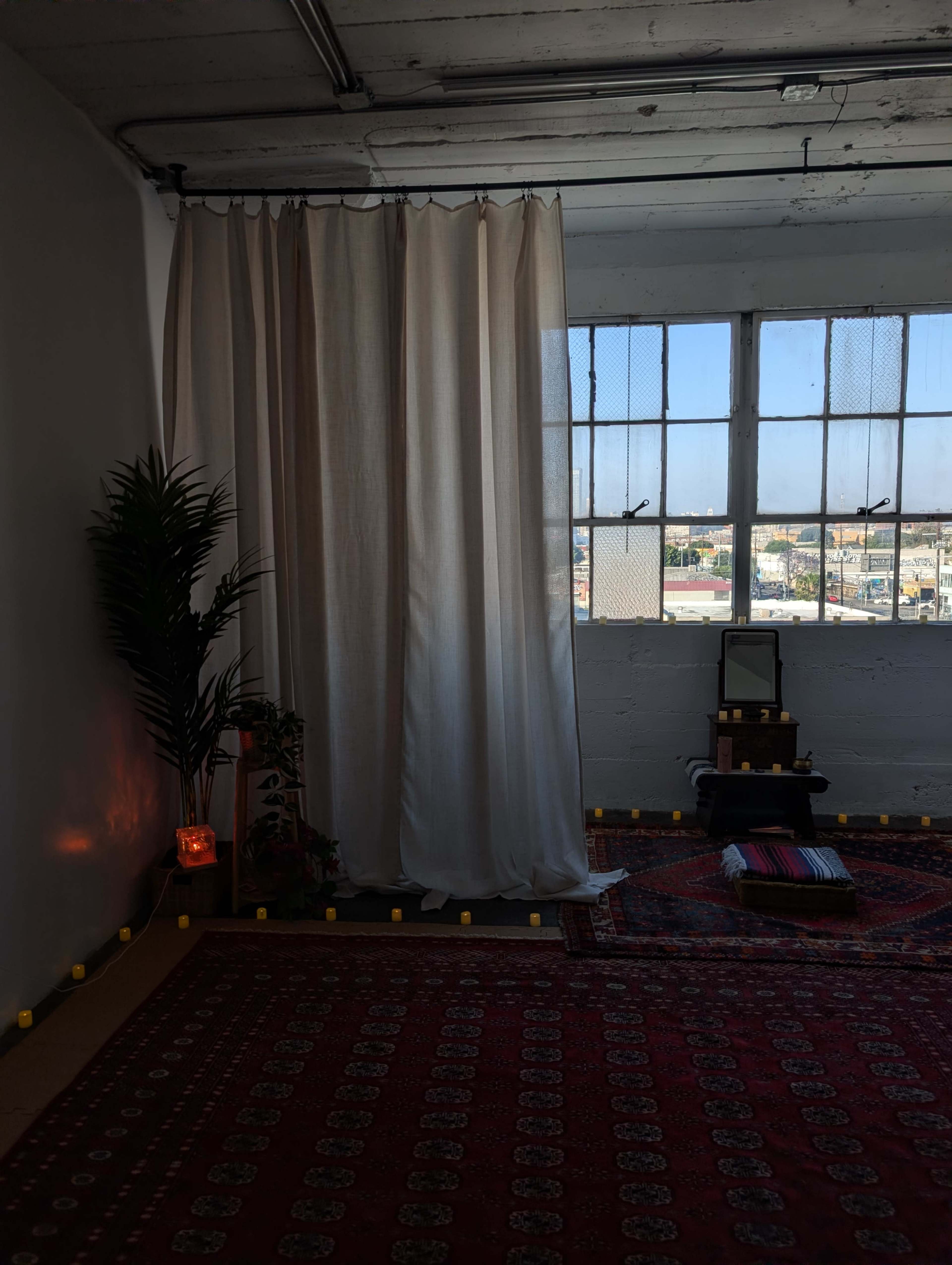 A bright room features a curtain, a potted plant, a mirror, and a patterned rug, with candles arranged along the floor near a window.
