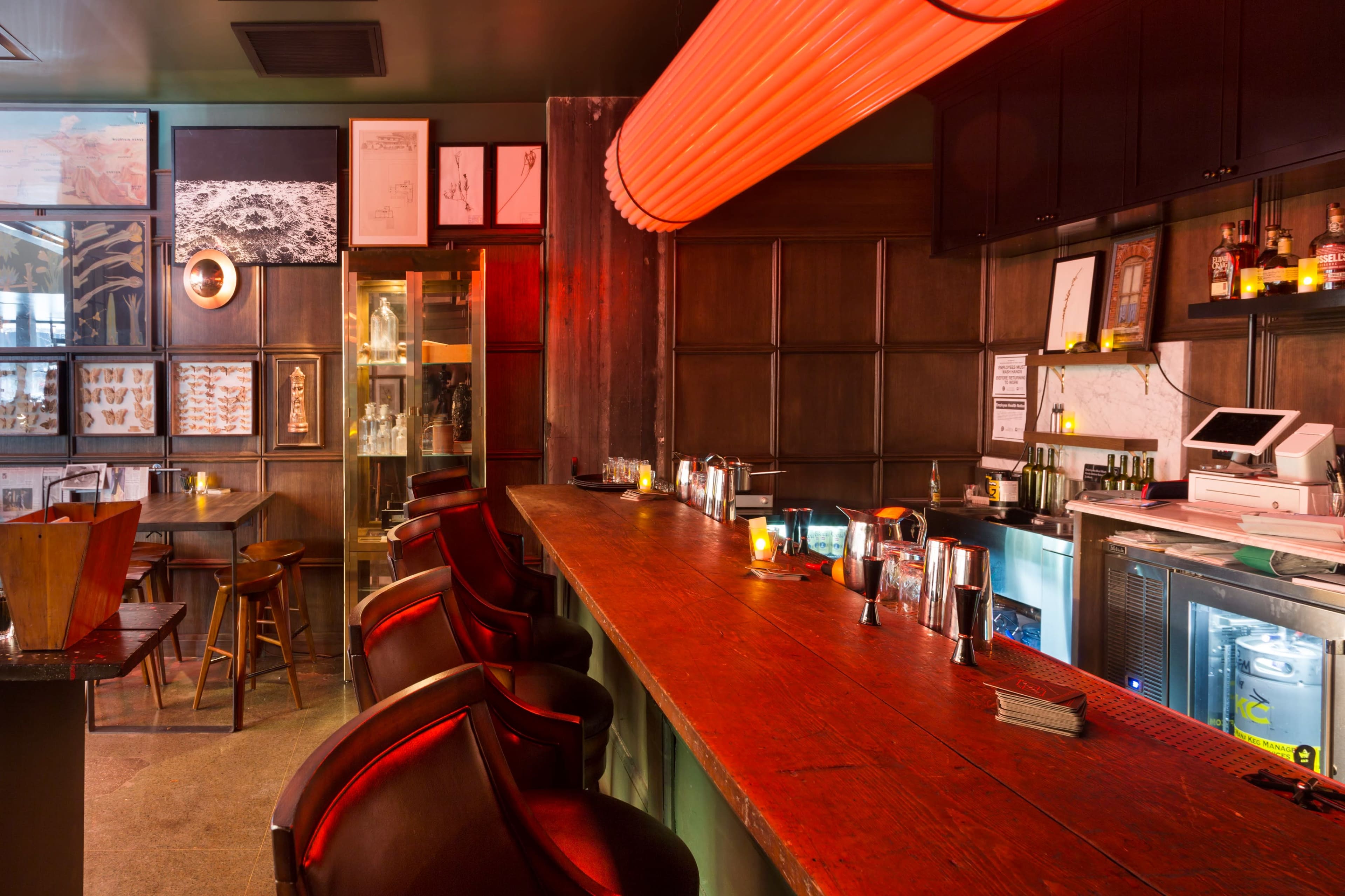 The image shows a wooden bar with high-backed chairs, illuminated by soft lighting, surrounded by framed artwork and shelves displaying bottles.