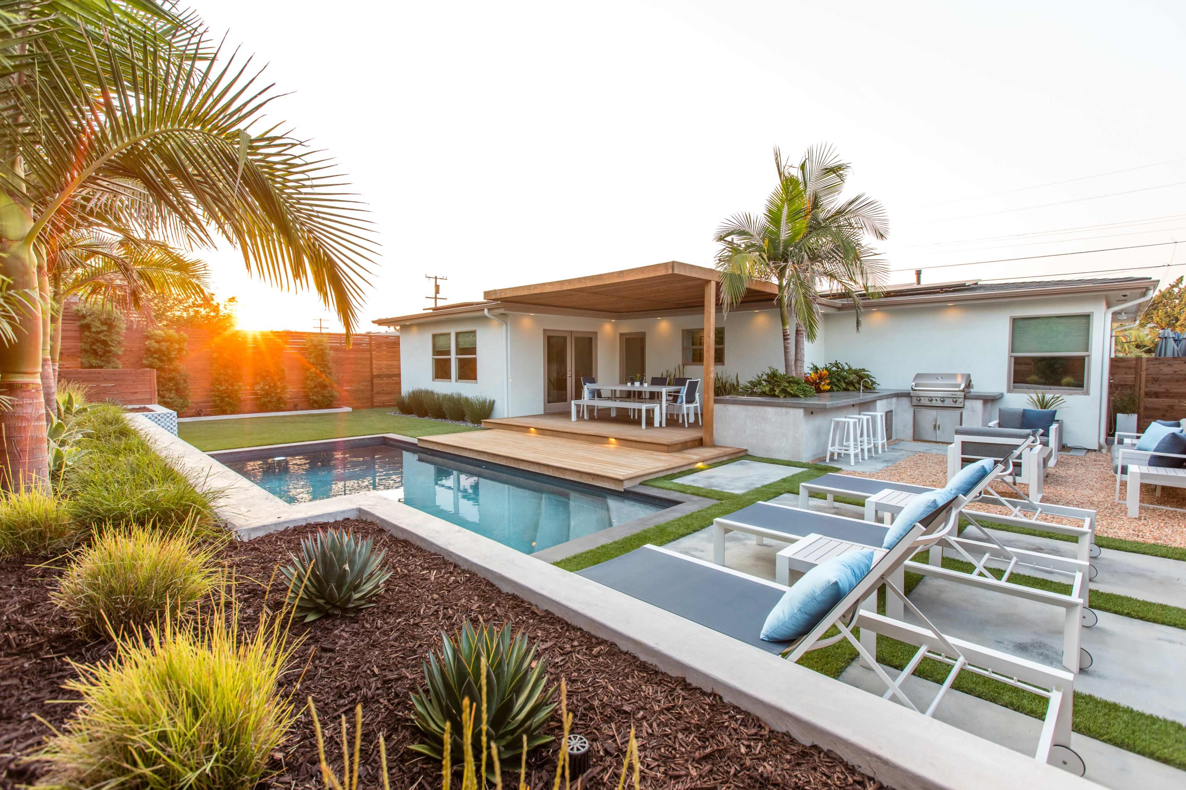 A modern backyard with a swimming pool, wooden deck, and seating area, surrounded by landscaping and palm trees at sunset.