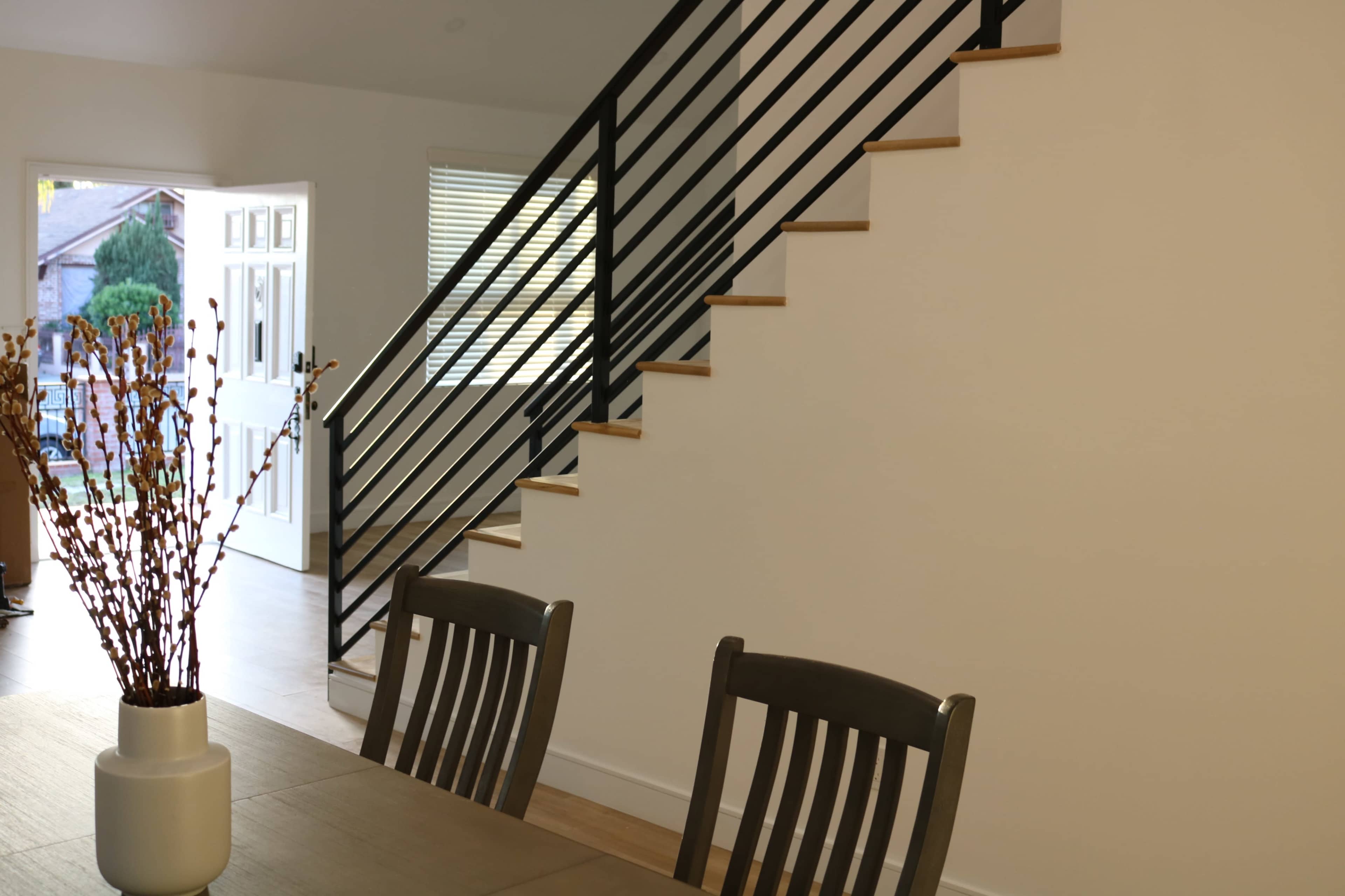 A staircase with black railings ascends along a white wall, while a modern dining table and chairs are positioned in the foreground.