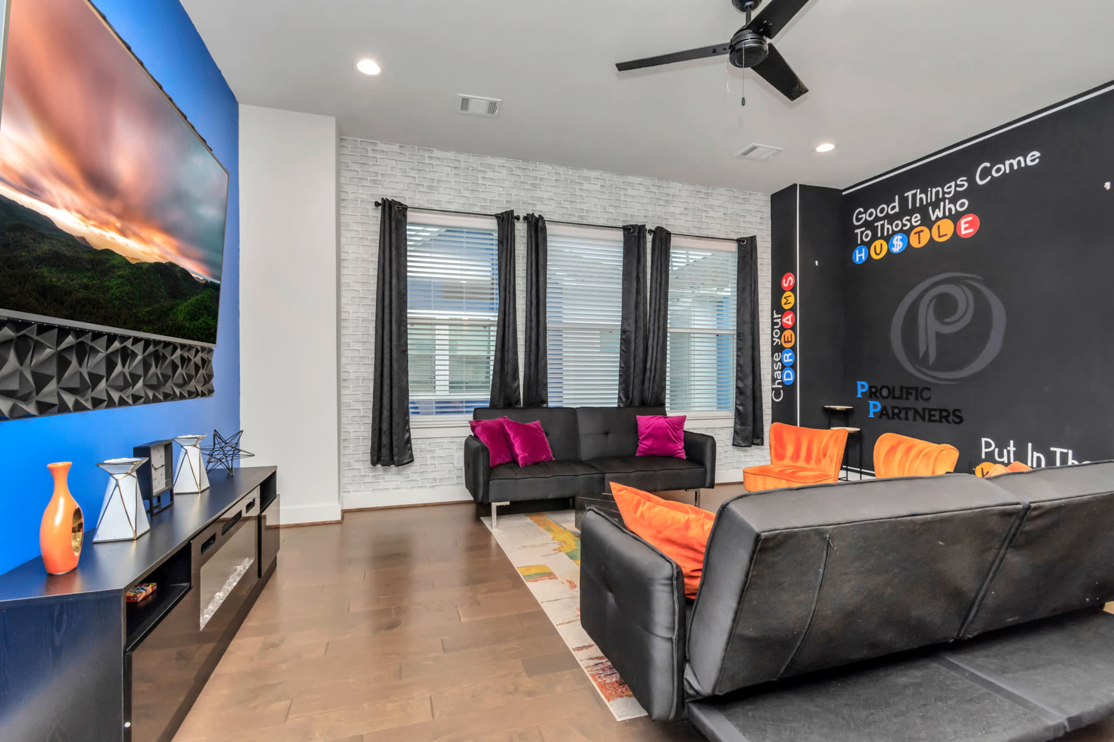 The image shows a modern living room with a large television, a black sofa set, colorful cushions, and a wall featuring motivational phrases.