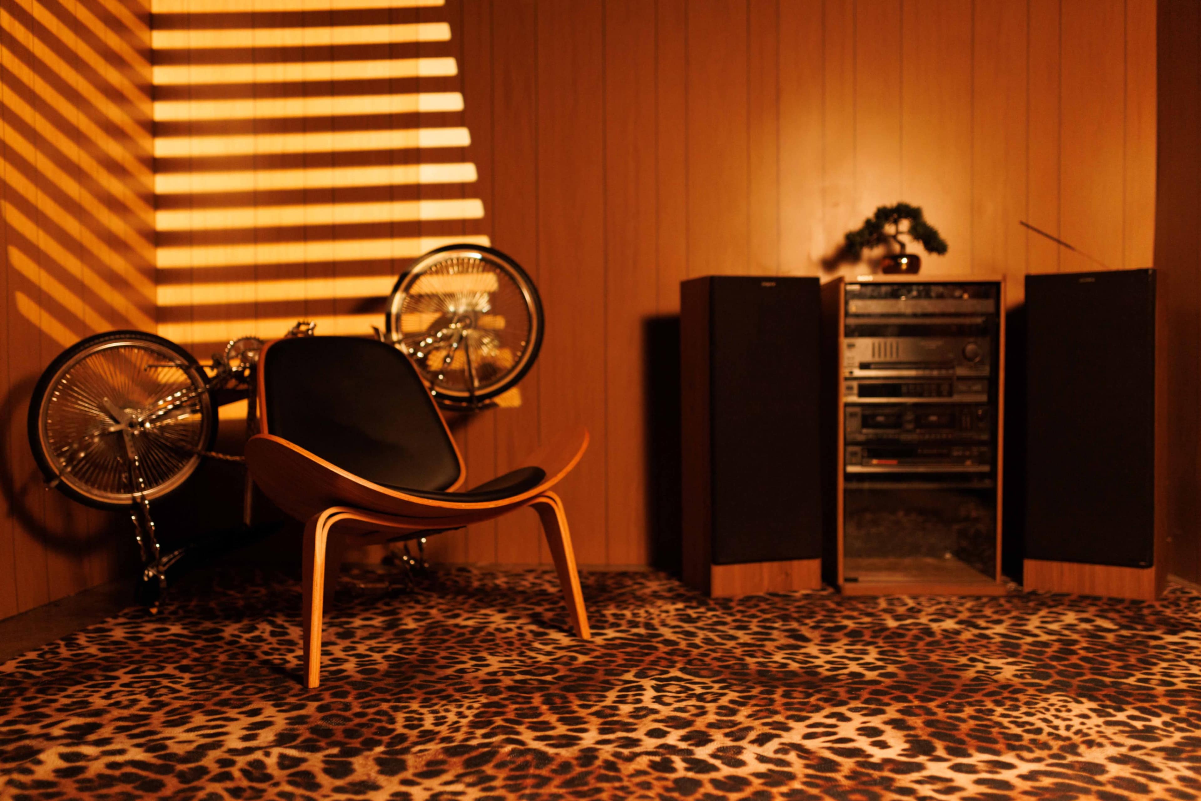 The image shows a retro-inspired interior with a sleek chair, a bicycle against the wall, and a stereo system flanked by tall speakers on a leopard-print rug.