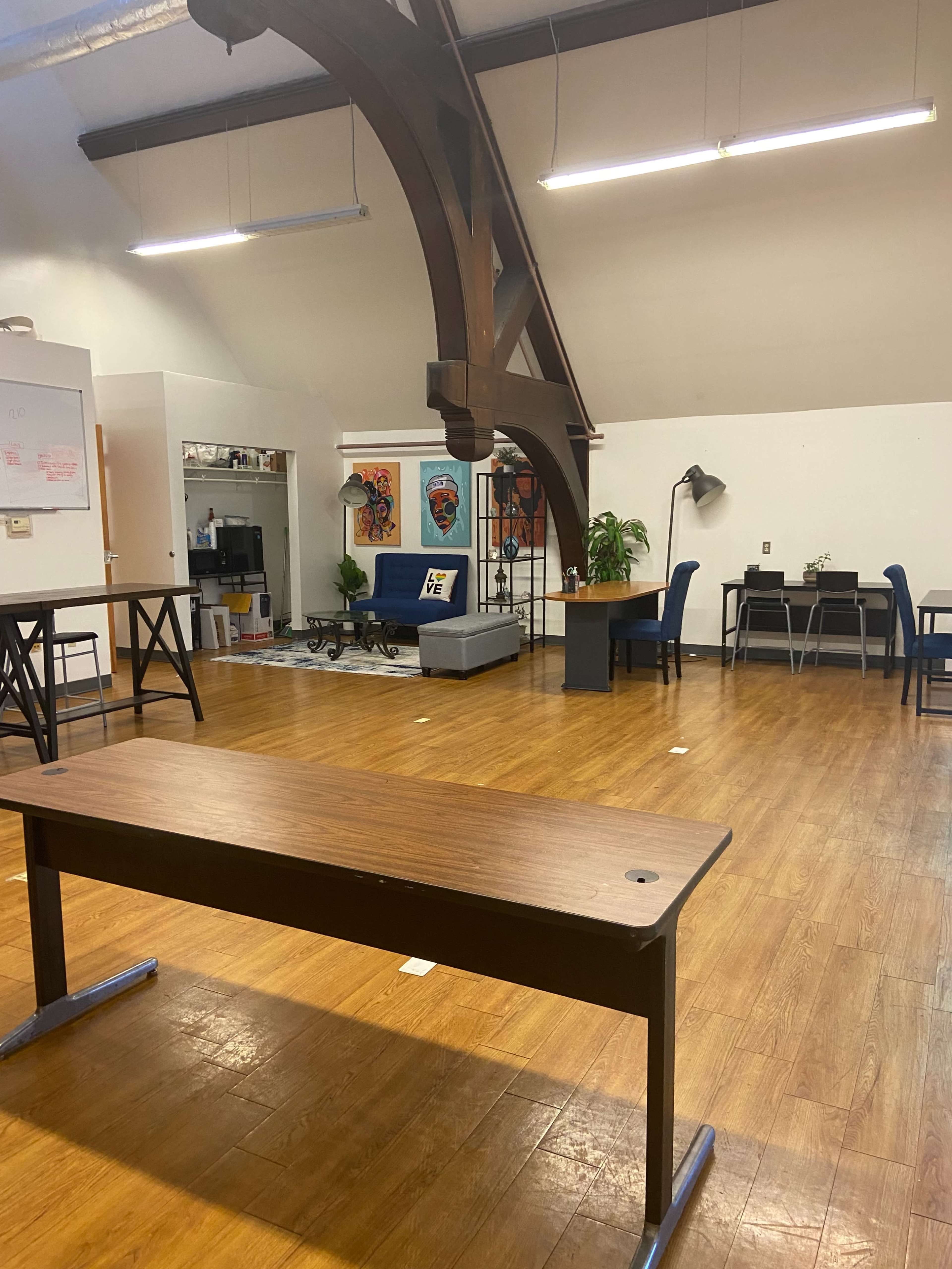 A spacious room with wooden flooring, featuring several tables and chairs, a sofa, and large beams on the ceiling.
