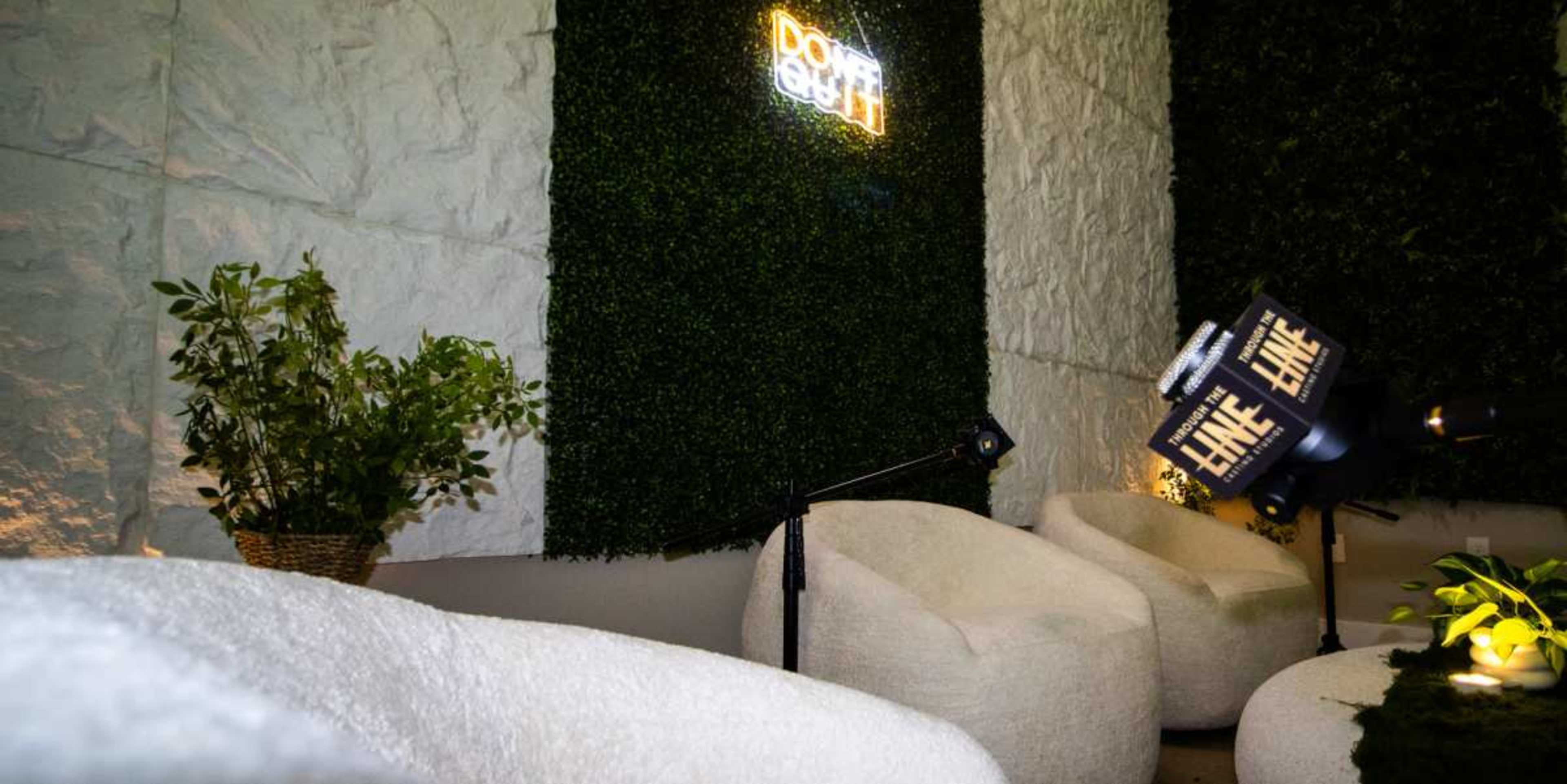 The image shows a cozy lounge area featuring two plush chairs, a plant, and a neon sign on a textured wall covered in greenery.