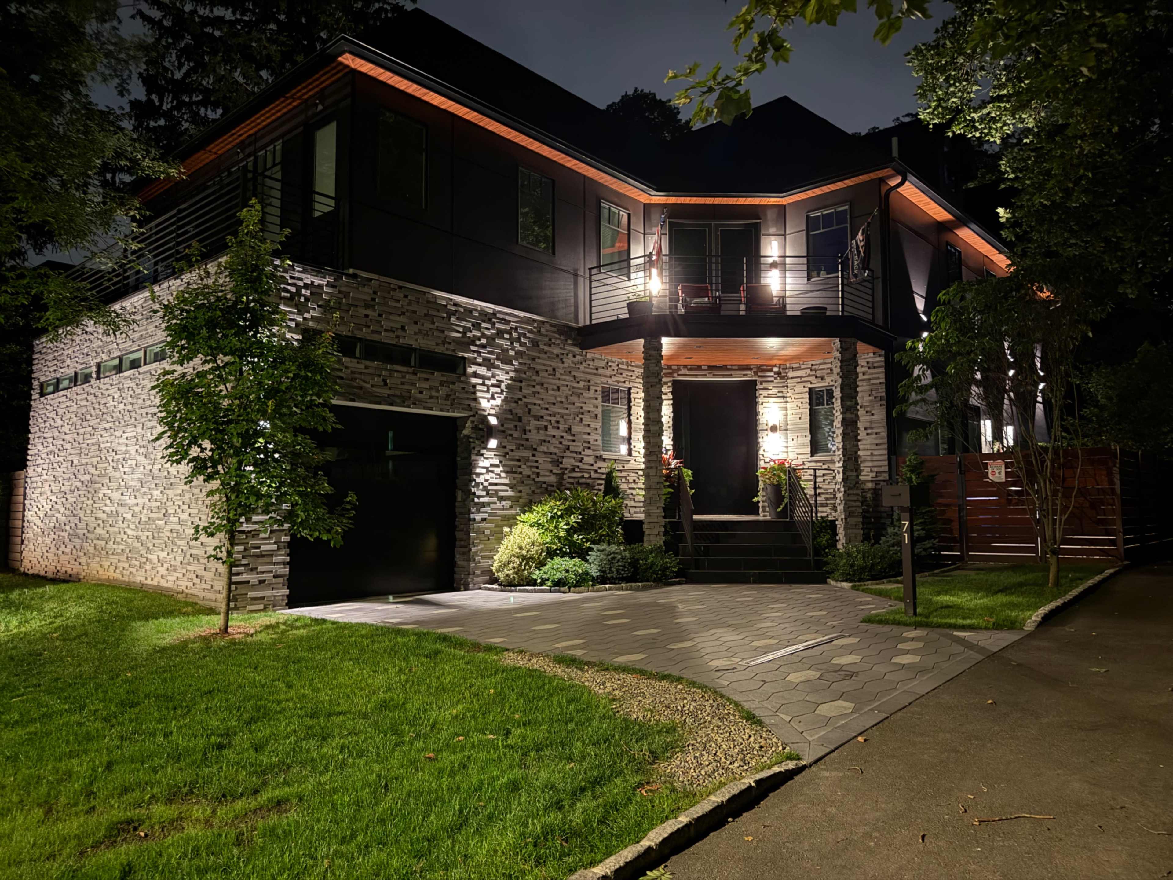 A modern house with stone and sleek black exterior features landscaping and outdoor lighting, set against a nighttime backdrop.