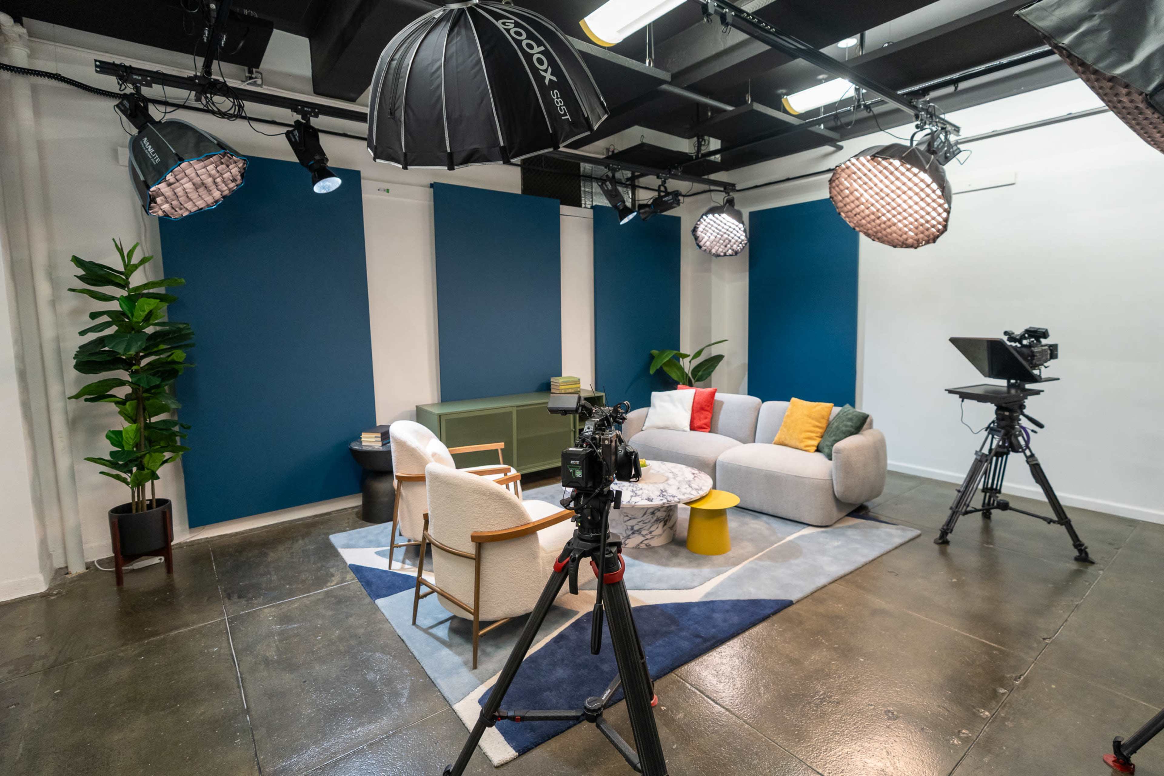 The image shows a television studio setup featuring a modern sofa, chairs, a coffee table, and multiple cameras under large lighting equipment.
