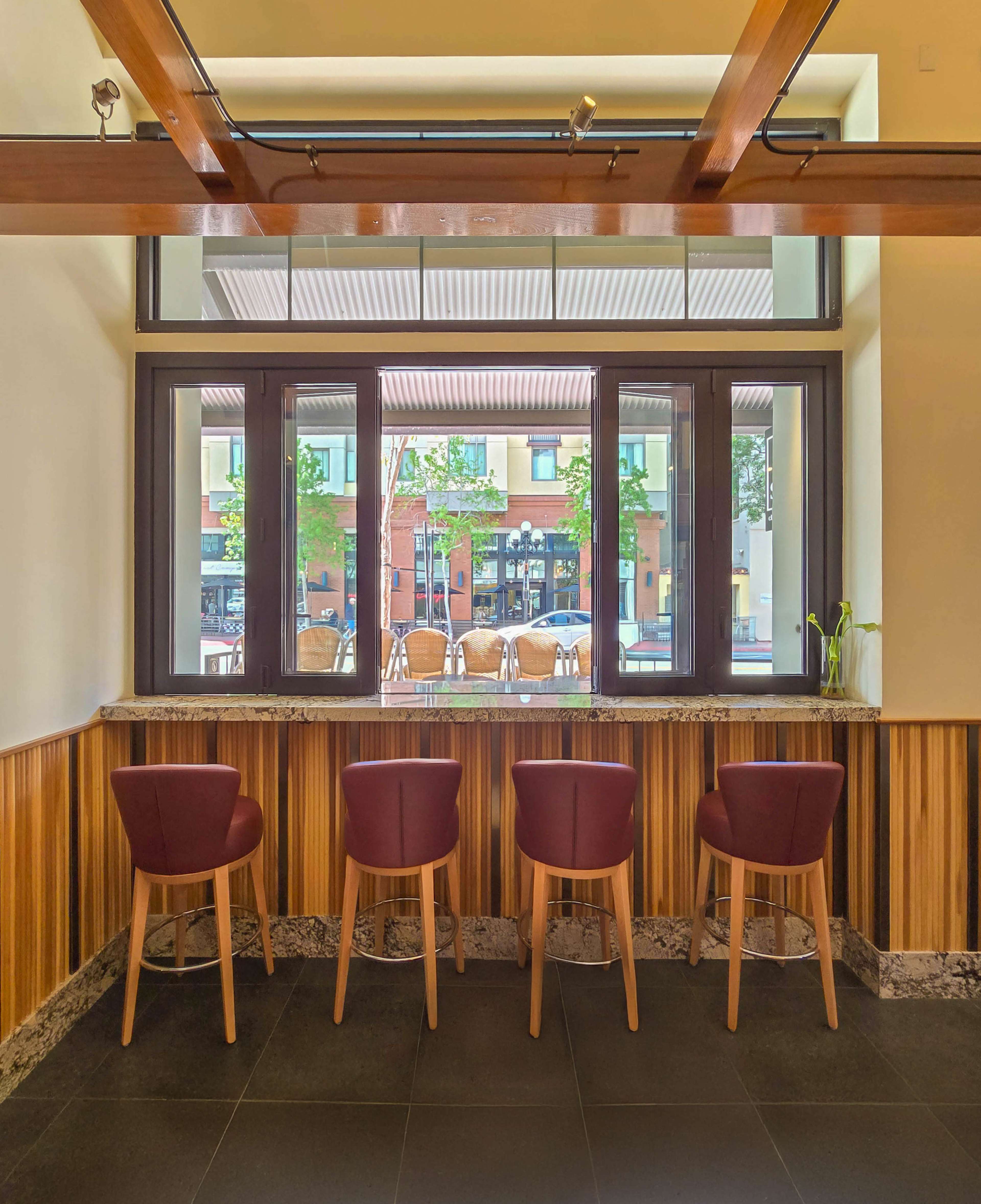 The image shows a modern interior with four maroon bar stools arranged in front of a window overlooking an outdoor seating area.