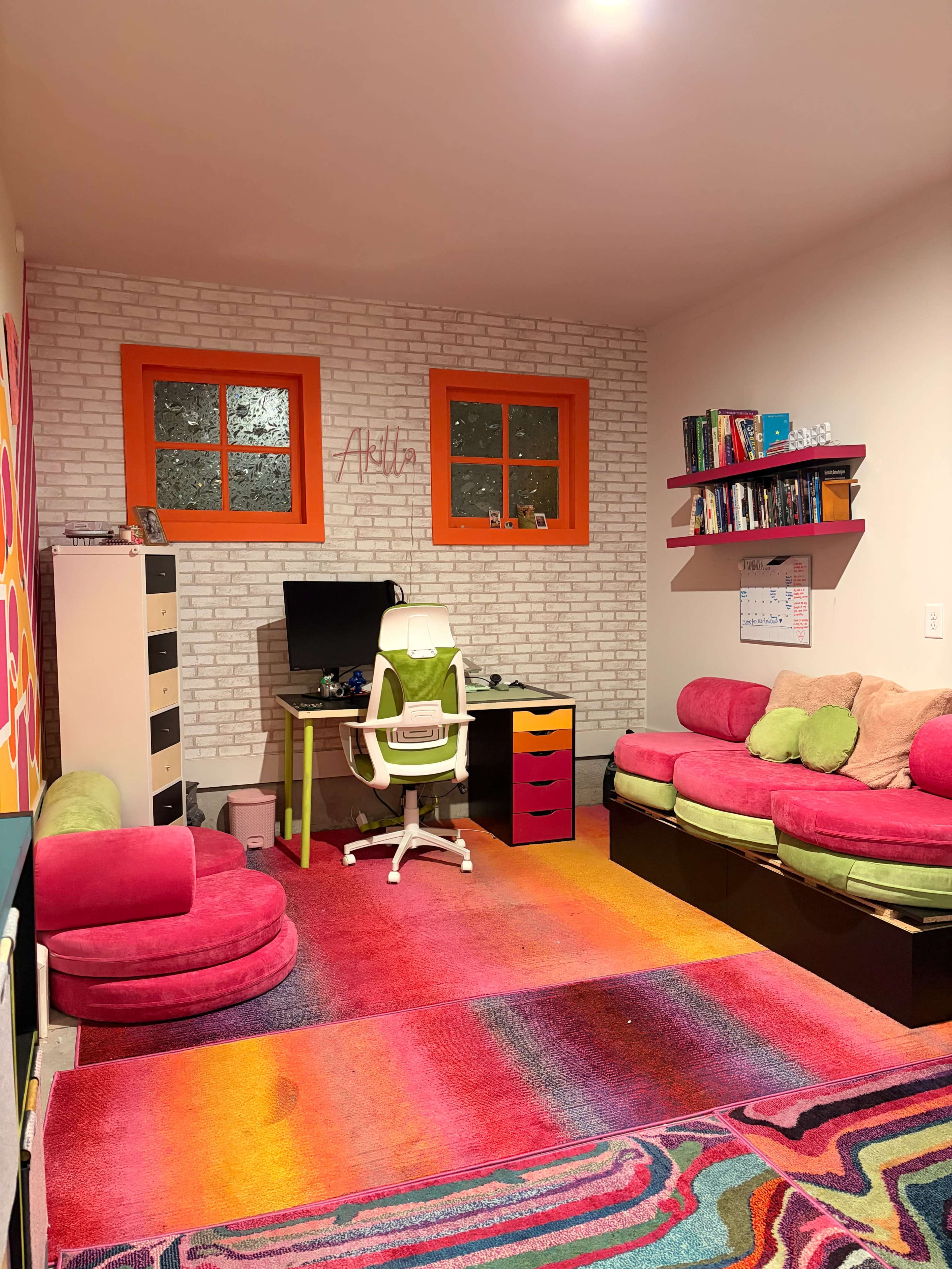 The room features a colorful design with a large area rug, a desk with a computer, and seating made up of pink and green cushions.