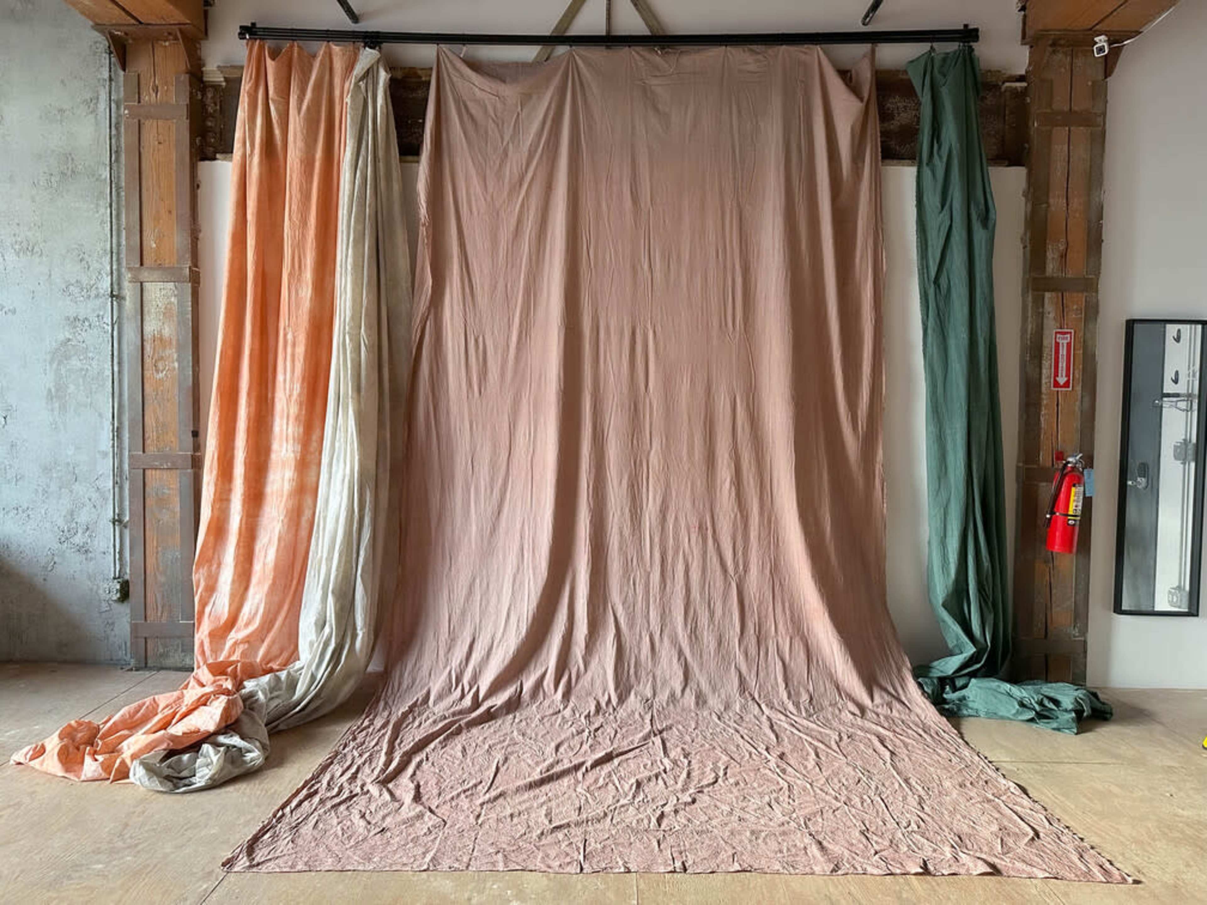 The image features a backdrop setup with large, wrinkled fabric drapes in shades of orange, cream, and green hanging vertically against a plain wall.