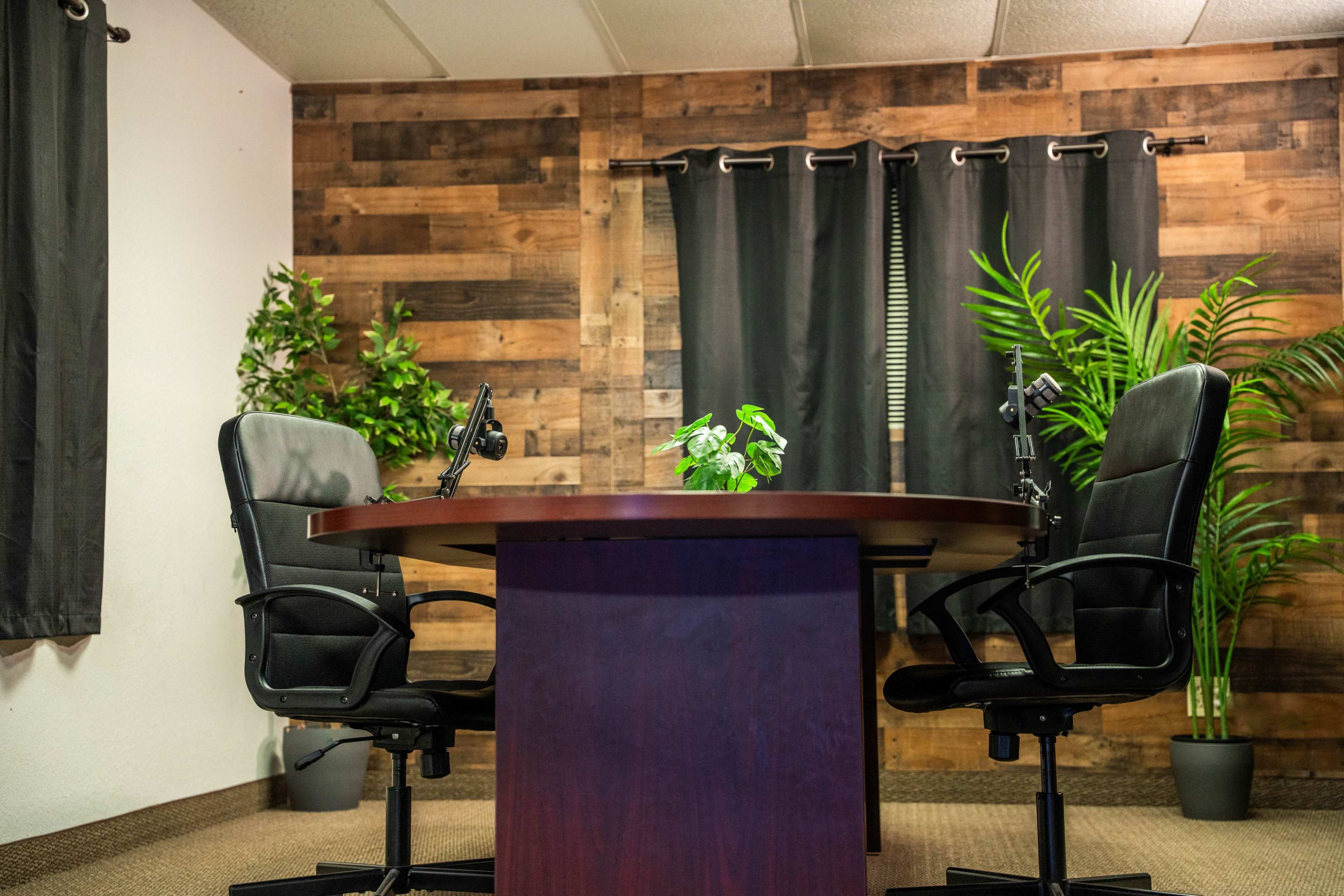 The image shows a small conference room with a round table, two rolling chairs, and plants near the walls.